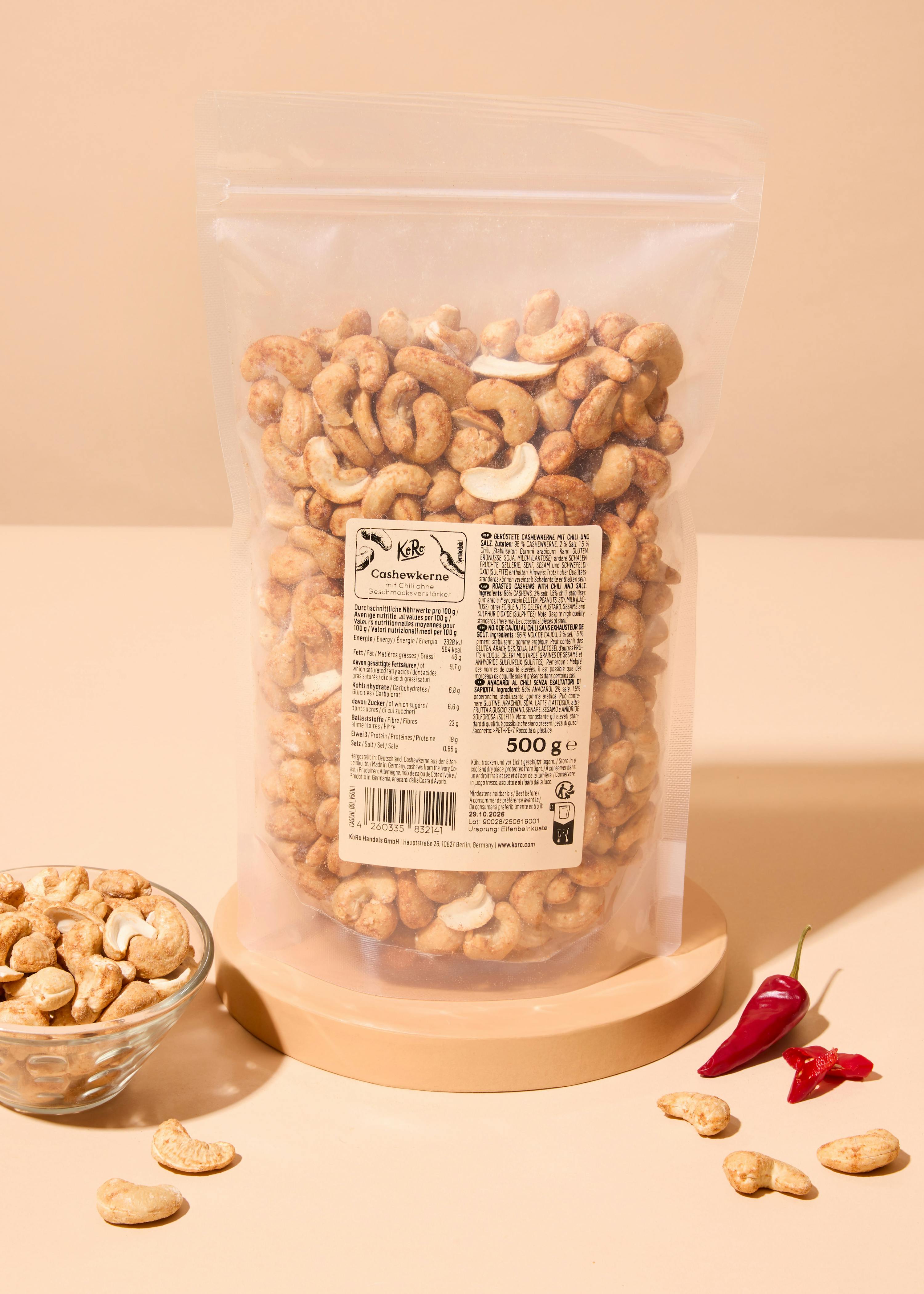 a clear plastic bag of roasted cashews with chili and salt sits on a wooden stand next to a small glass bowl of cashews and two red chili peppers.