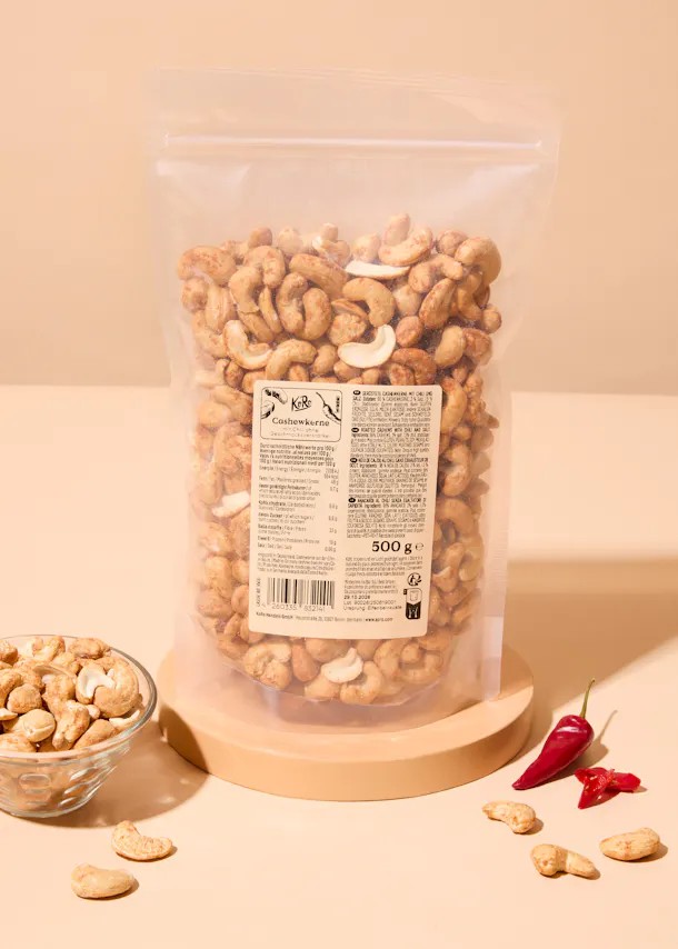 un sac transparent rempli de noix de cajou grillées, posé sur un support en bois clair, avec un petit bol de noix de cajou et des piments rouges à côté.