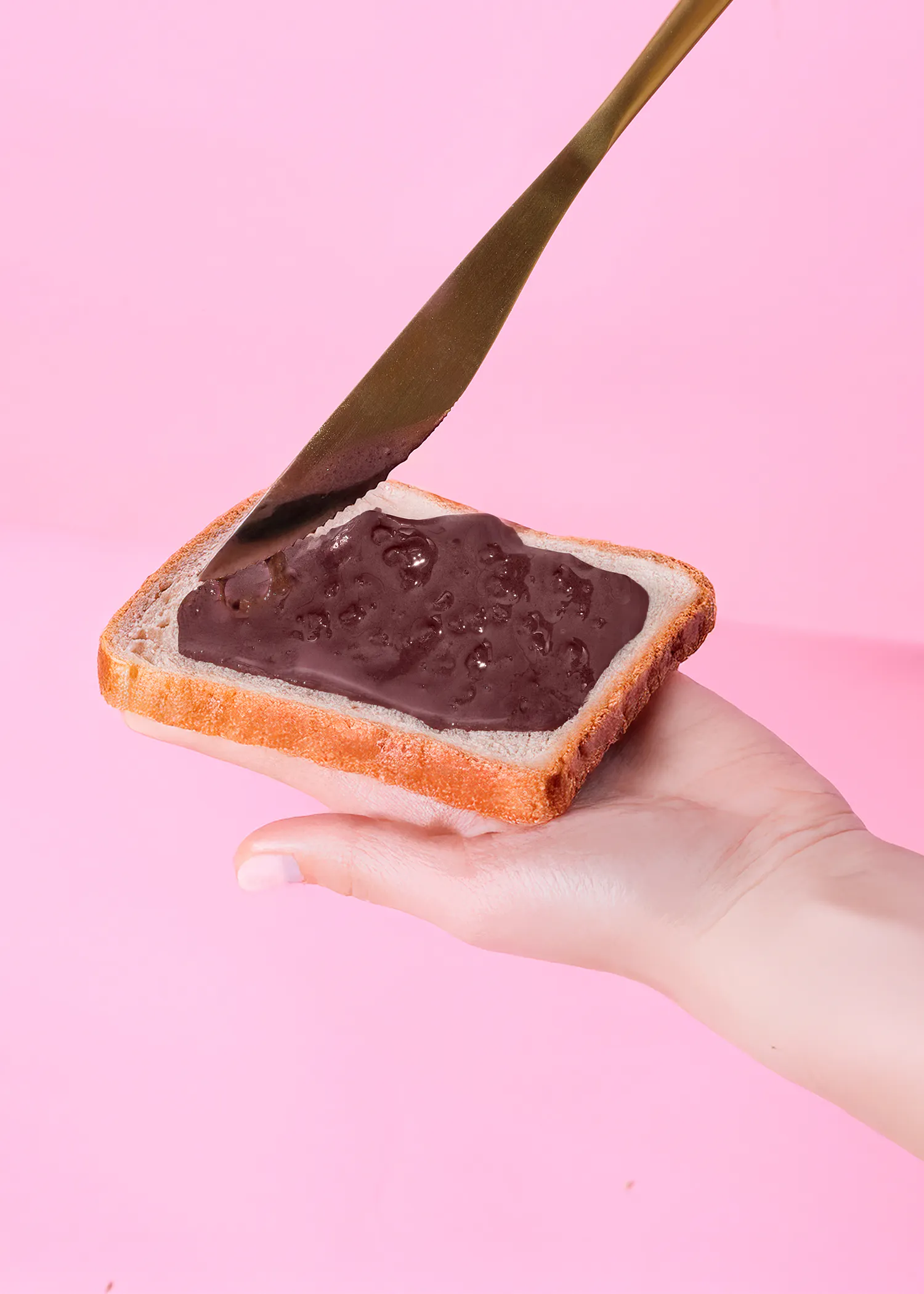 une main tient une tranche de pain blanc avec de la pâte à tartiner au chocolat, étalée avec un couteau sur un fond rose.