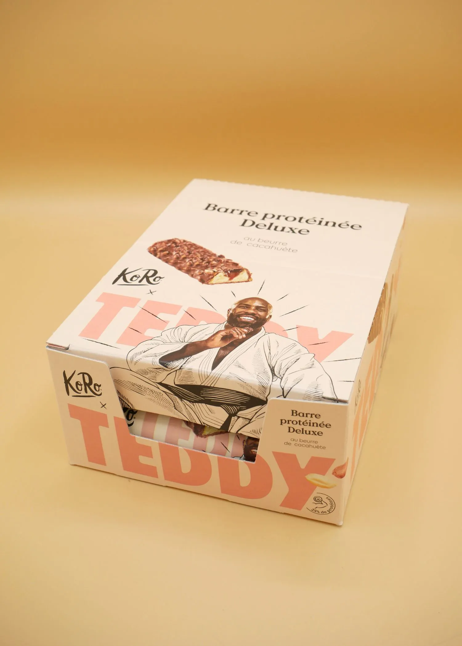 eine schachtel koro deluxe proteinriegel mit erdnussbuttergeschmack, verziert mit einer illustration eines lächelnden mannes in einem judo-gi und dem schriftzug 'teddy'.