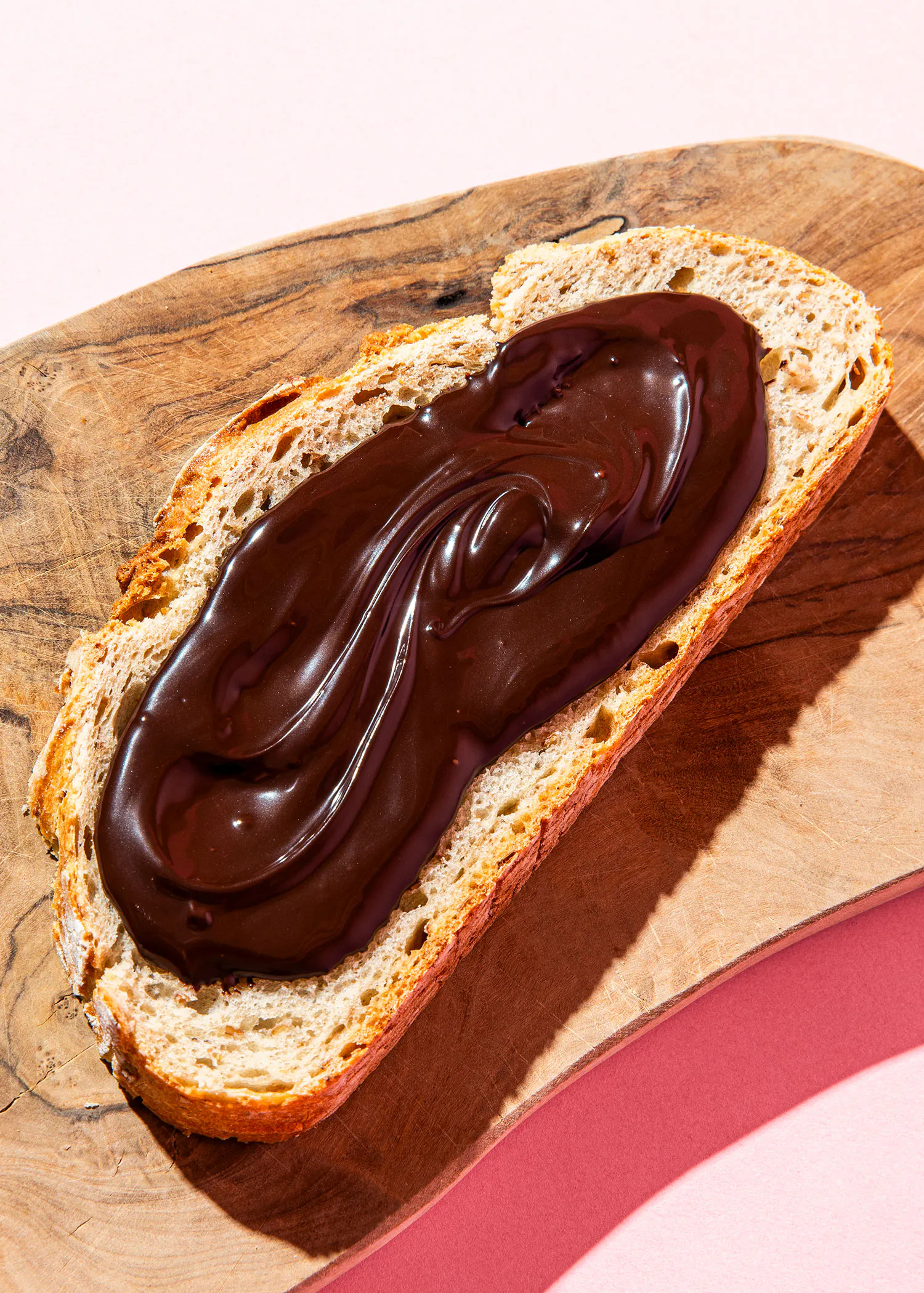 eine scheibe rustikales brot mit einer dicken, glänzenden schicht dunkler schokoladencreme auf einem holzbrett.