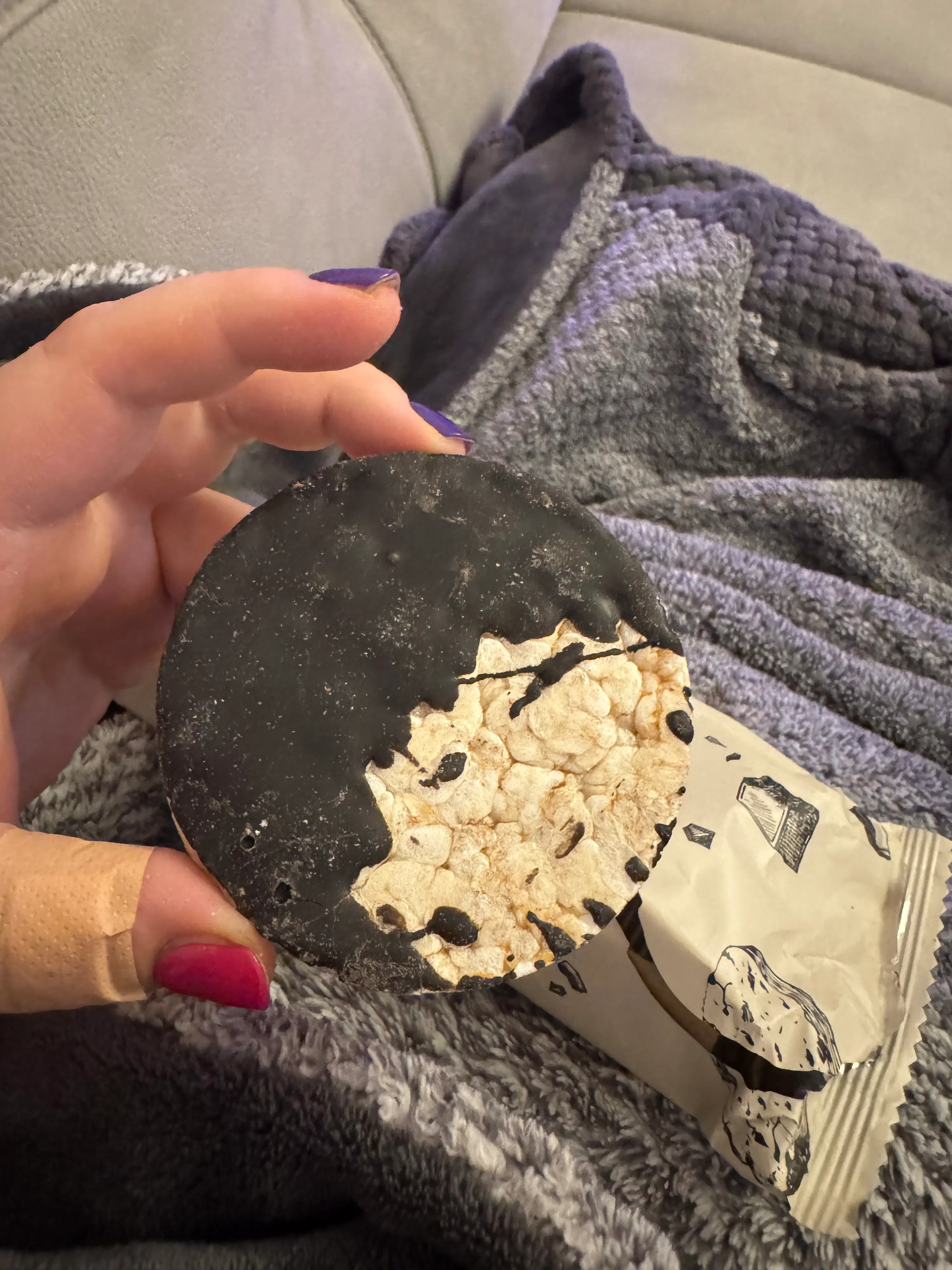 a hand holds a round rice cake partially covered in dark chocolate, with a fuzzy blanket and snack packaging in the background.