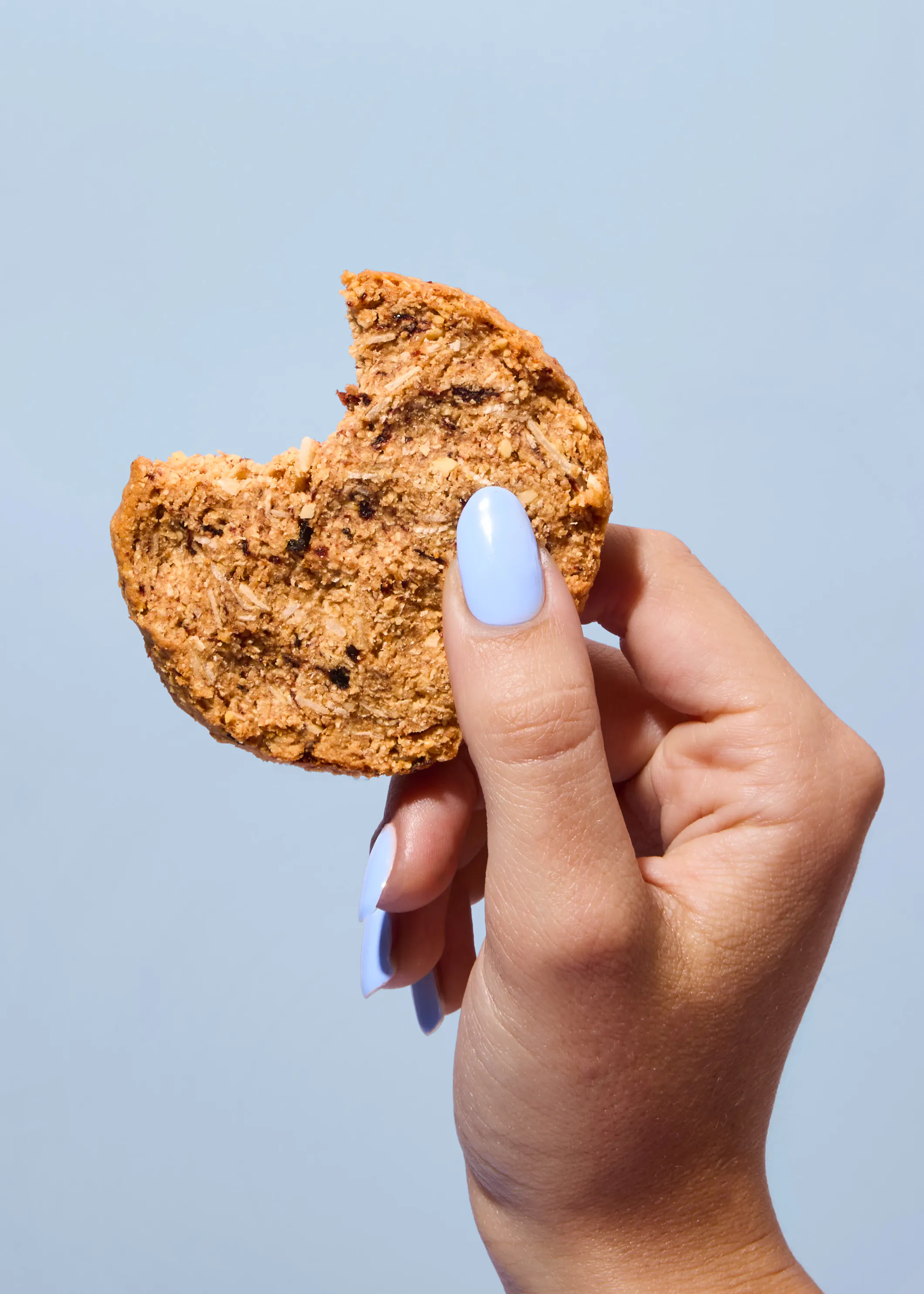 une main aux ongles vernis en bleu clair tient un biscuit aux céréales entamé sur un fond bleu uni.