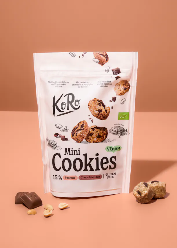 una bolsa blanca de mini galletas KoRo veganas y sin gluten con trozos de chocolate y cacahuetes, rodeada de galletas sueltas, trozos de chocolate y cacahuetes sobre un fondo melocotón.