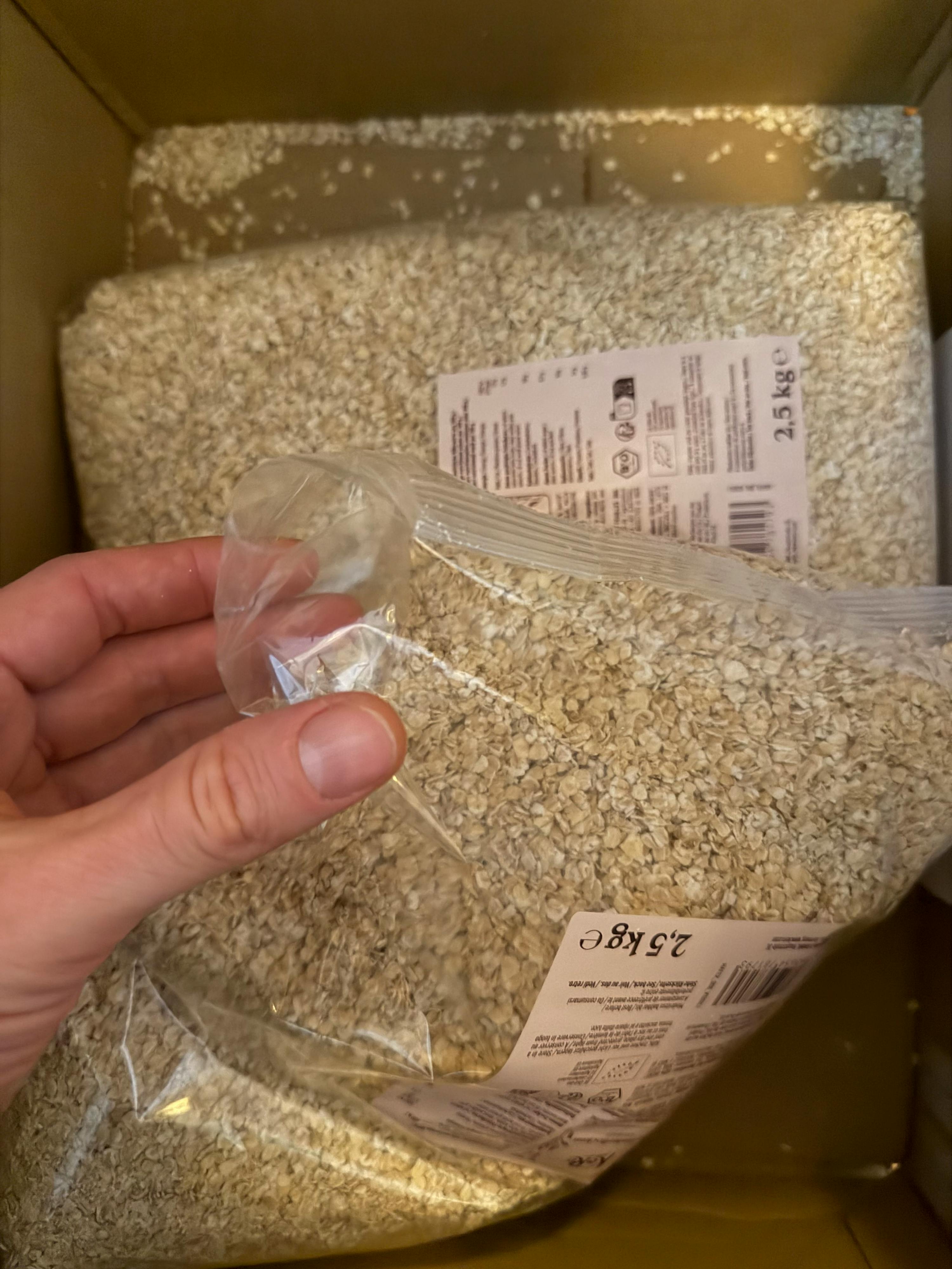 main tenant un sac transparent de flocons d'avoine de 2,5 kg, avec un autre sac similaire en arrière-plan dans une boîte en carton.