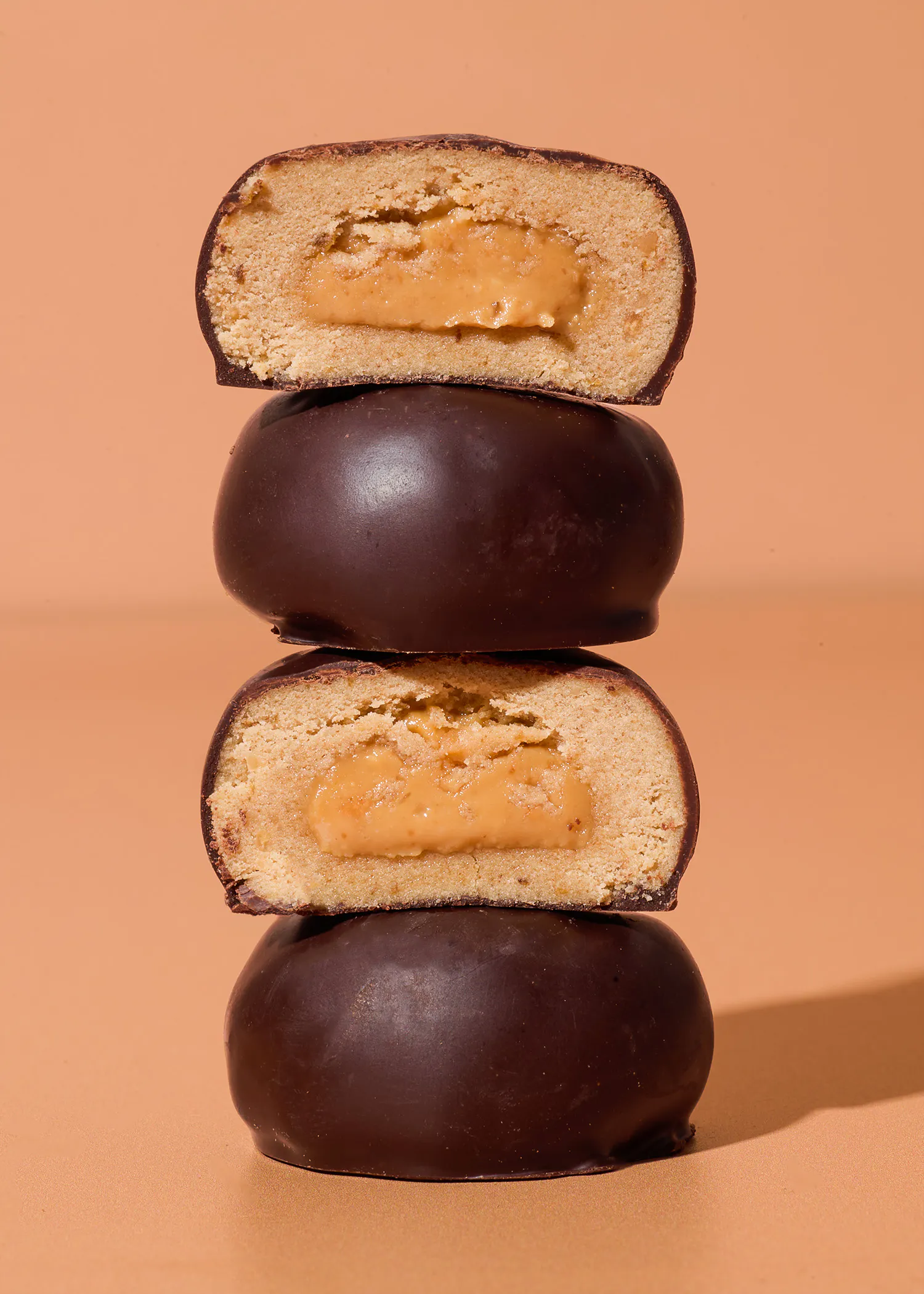 una pila di quattro dolcetti ricoperti di cioccolato fondente, di cui due tagliati a metà per mostrare il ripieno cremoso color caramello su uno sfondo color pesca.