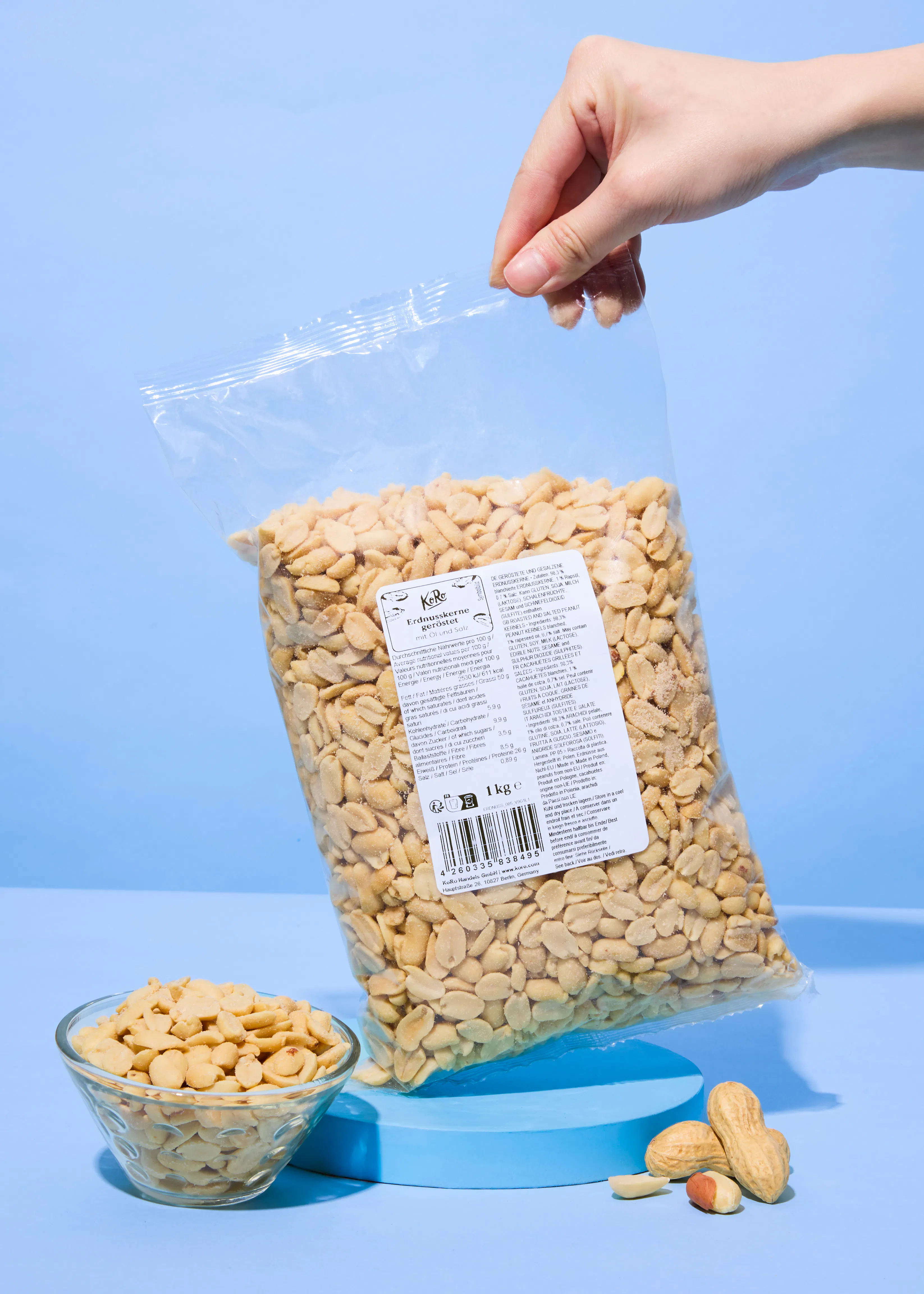 une main tient un grand sac transparent de 1 kg de cacahuètes grillées k&ro, avec un bol de cacahuètes et des cacahuètes éparpillées sur une surface bleue.