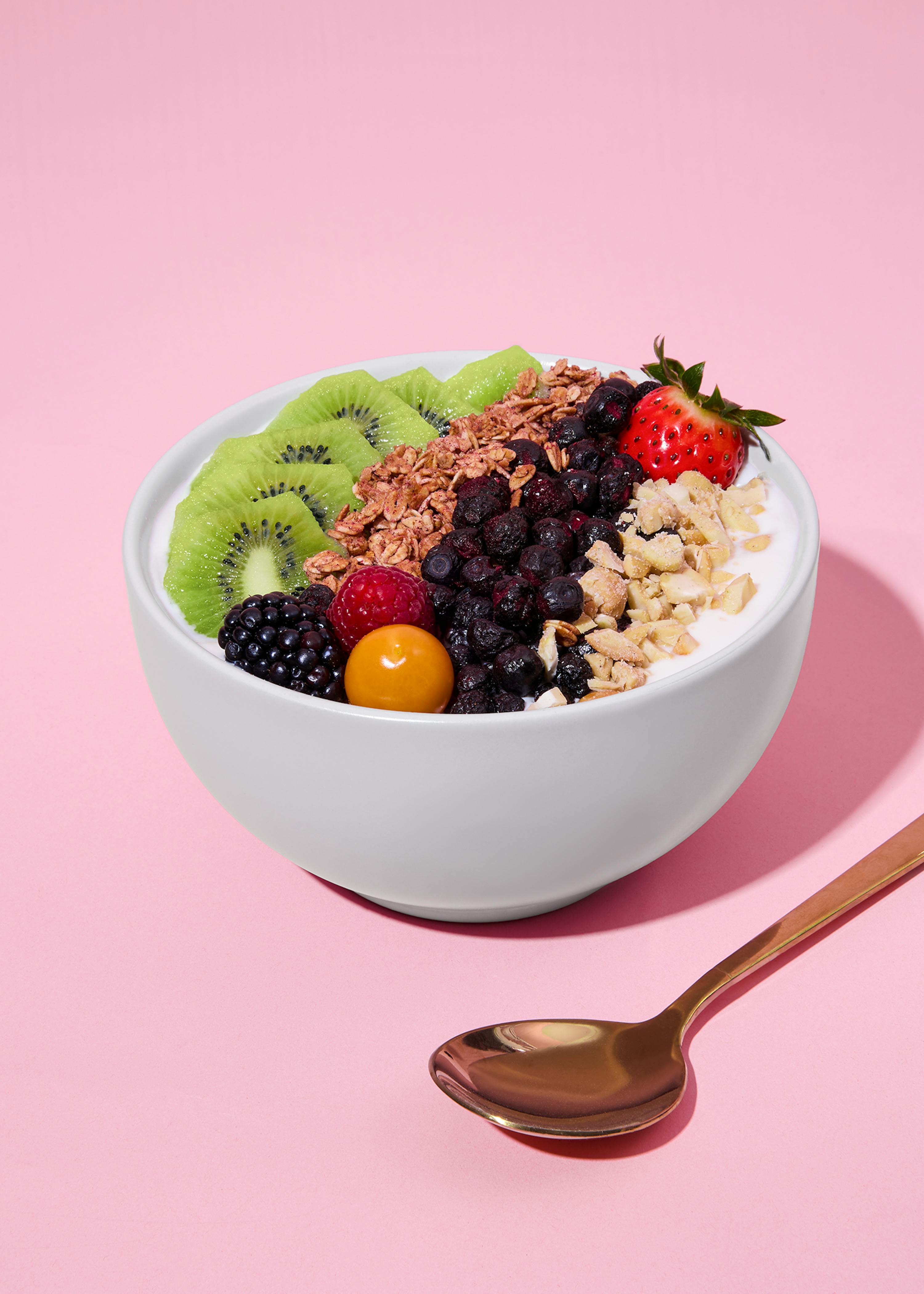 a white bowl of yogurt topped with sliced kiwi, granola, dried blueberries, a strawberry, chopped nuts, a blackberry, a raspberry, and a goldenberry, with a gold spoon on a pink background.