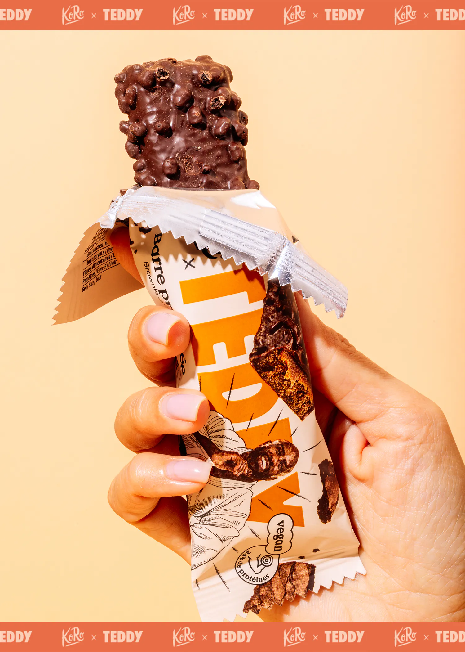 une main tient une barre protéinée au chocolat partiellement déballée, avec un enrobage croustillant et un emballage illustré d'un homme souriant et des mentions 'vegan' et 'protéines'.