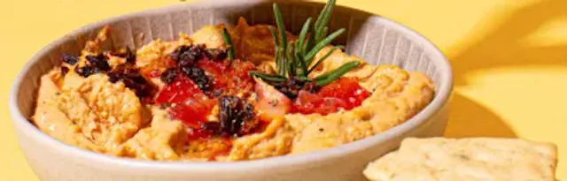 un tazón de hummus con tomates y romero junto a un trozo de galletas .