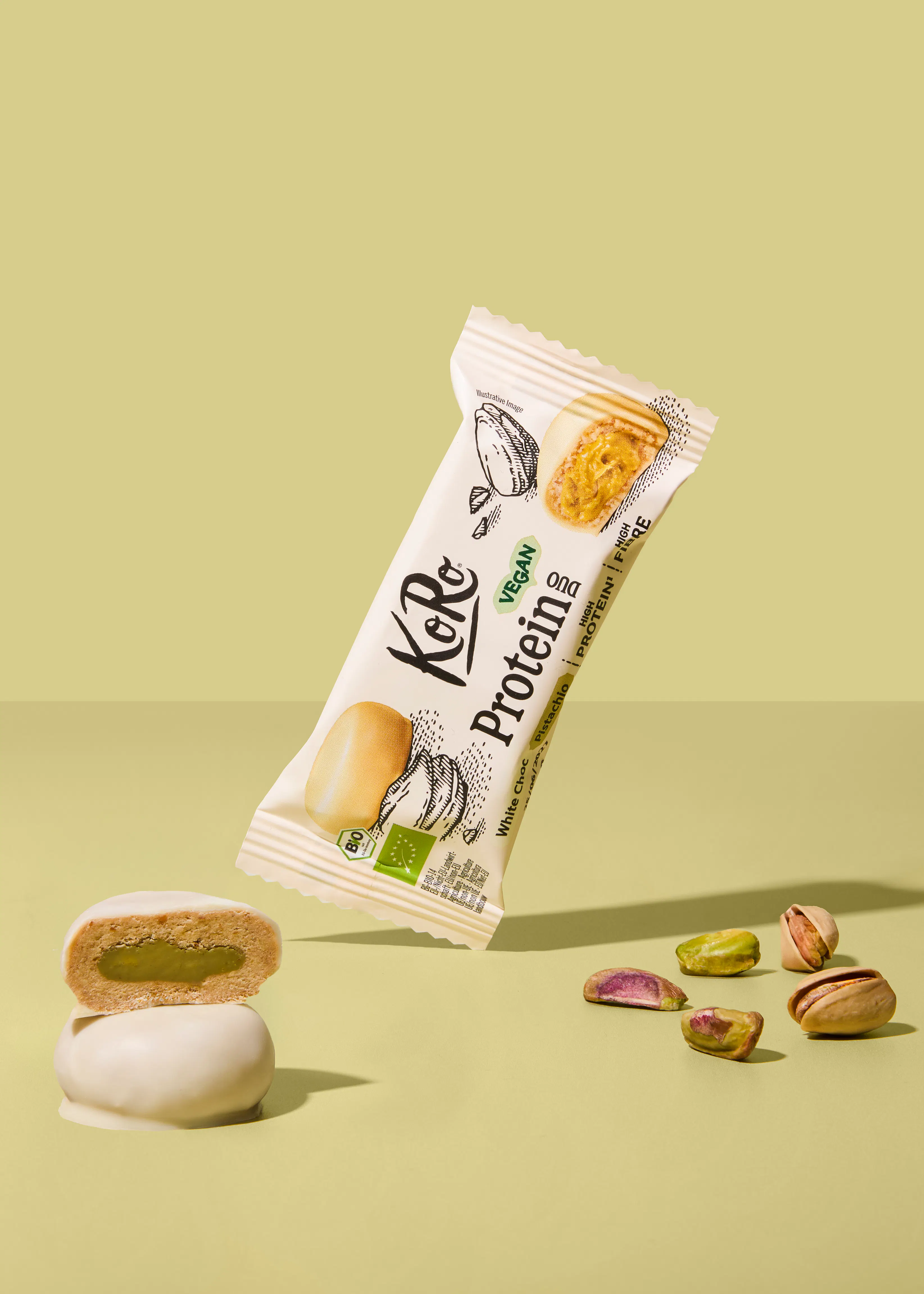 eine schwebende, weiße koro proteinriegelverpackung mit der aufschrift „white choc pistachio“ über zwei gestapelten, halbierten weißen schokoladensnacks mit grüner füllung und verstreuten pistazien auf hellgrünem hintergrund.