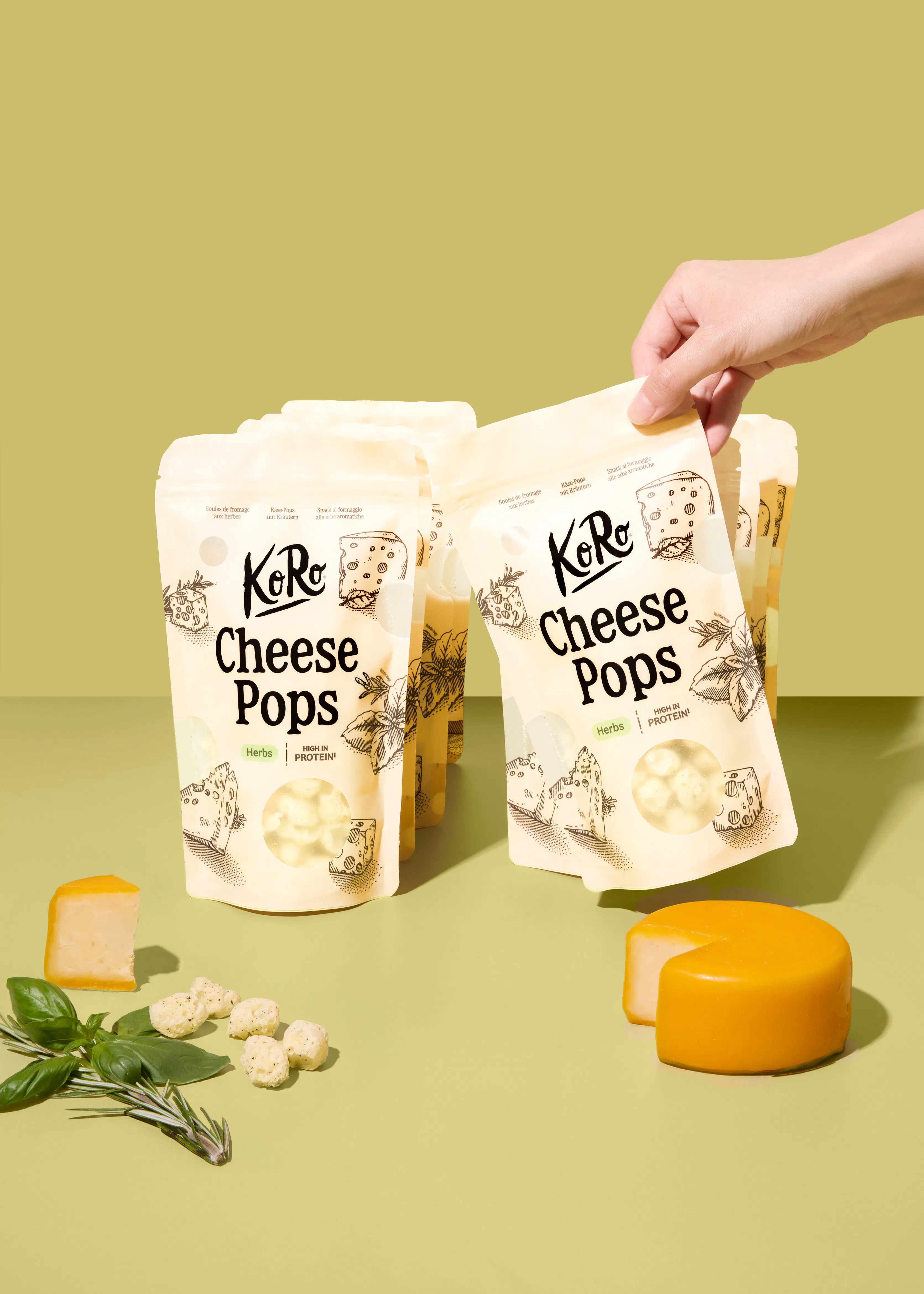 eine hand hält eine packung koro cheese pops vor weitere packungen, während auf dem tisch ein stück käse, ein kleiner käselaib und frische kräuter liegen.