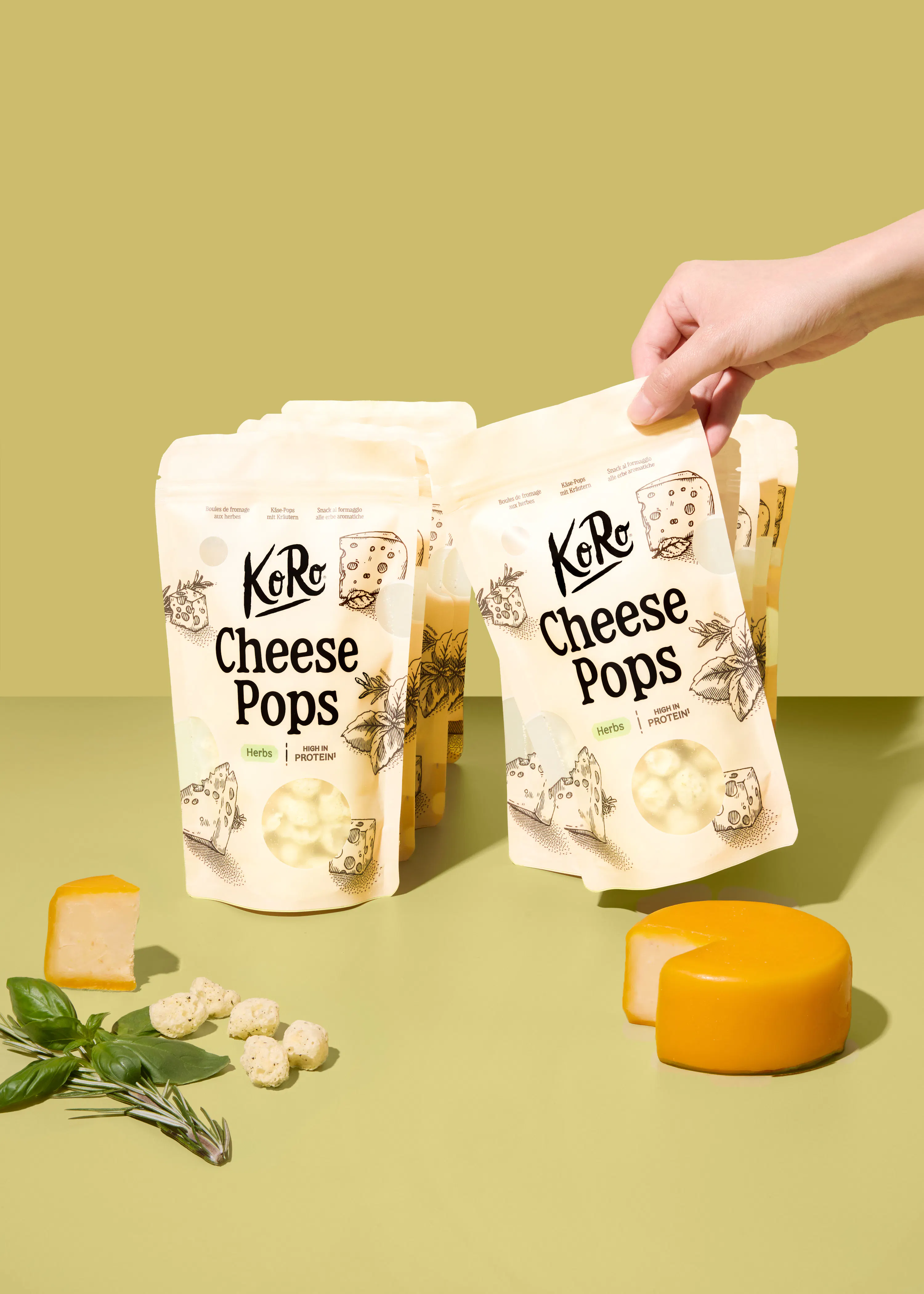 una mano tiene una confezione di koro cheese pops alle erbe su uno sfondo verde chiaro, circondata da pezzi di formaggio e rametti di basilico.