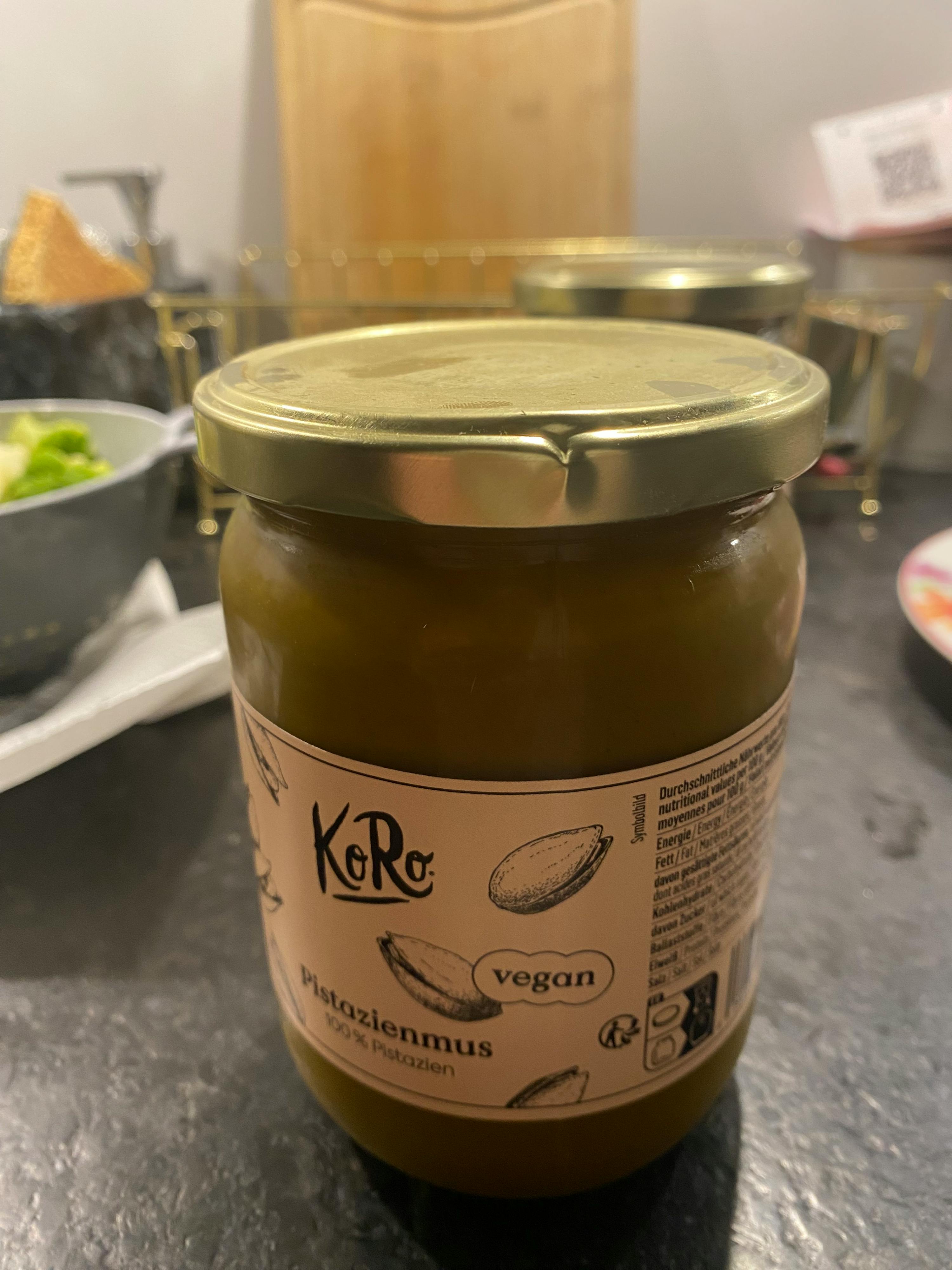 a jar of koro 100% pistachio butter with a gold lid and a vegan label, lying on a dark countertop.