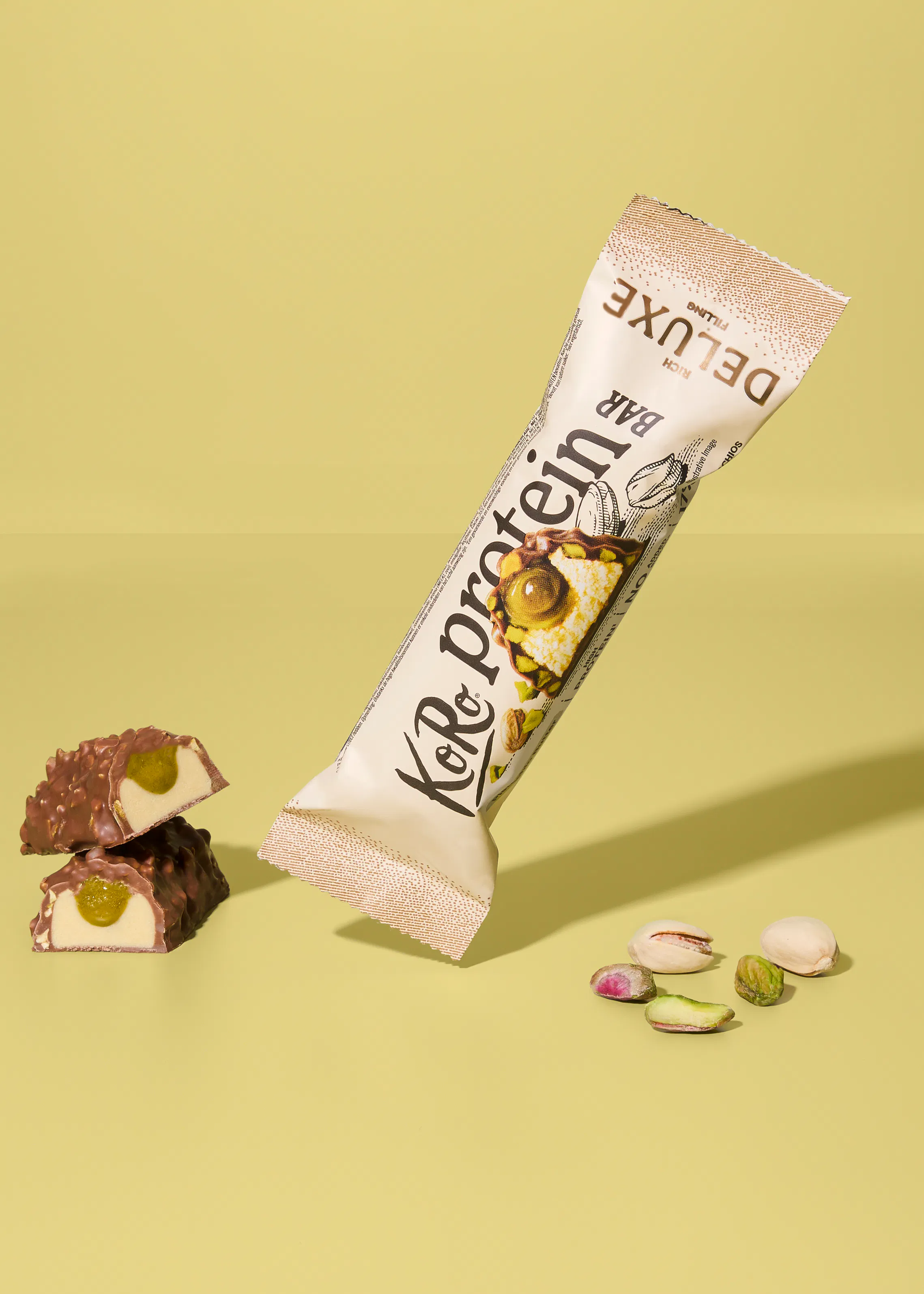 eine ko&protein deluxe bar in der verpackung, daneben zwei aufgeschnittene riegelhälften mit pistazienfüllung und mehrere lose pistazien auf hellgelbem hintergrund.