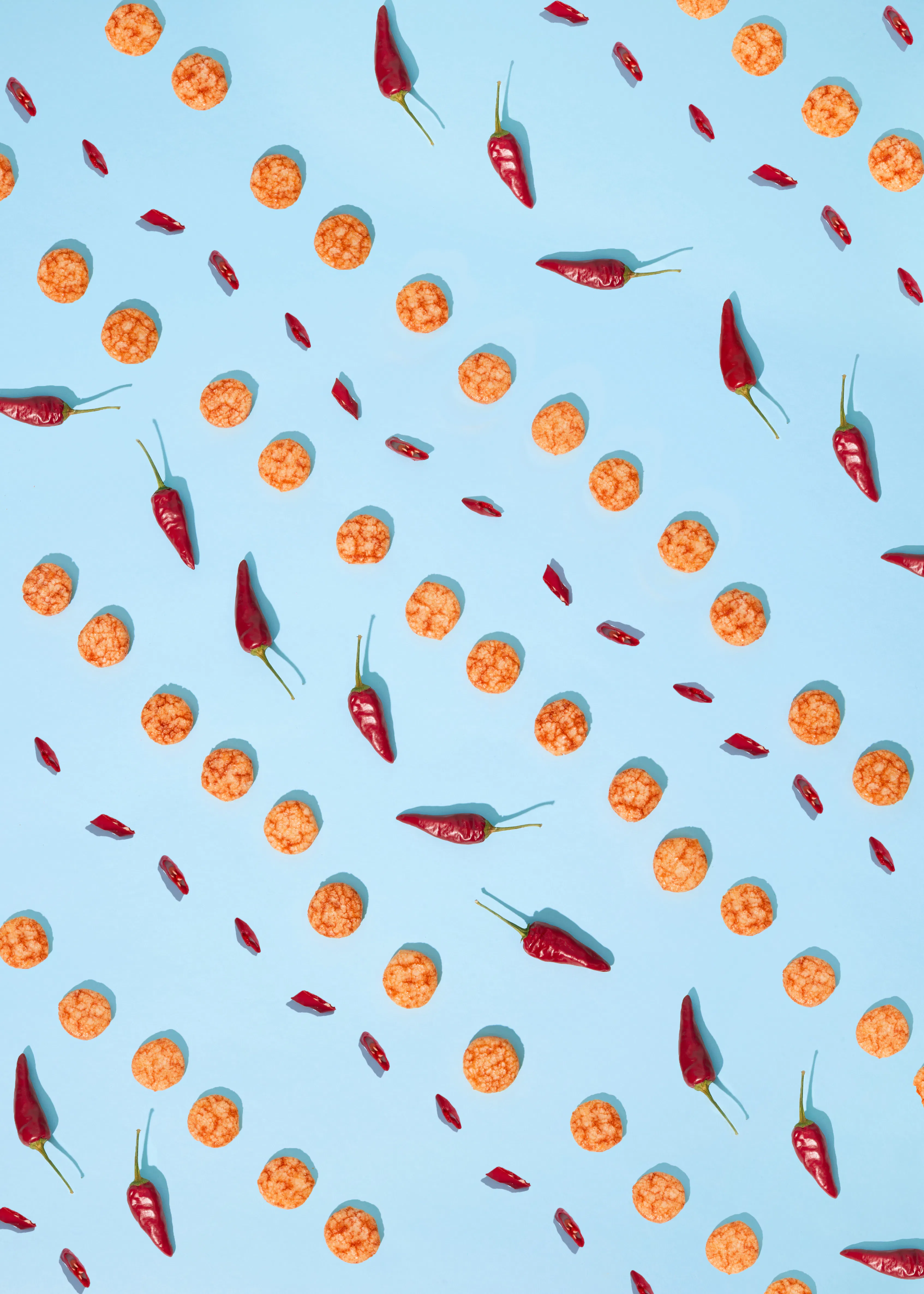 un motif de piments rouges entiers et coupés en rondelles, et de petits biscuits ronds orange, dispersés sur un fond bleu clair.
