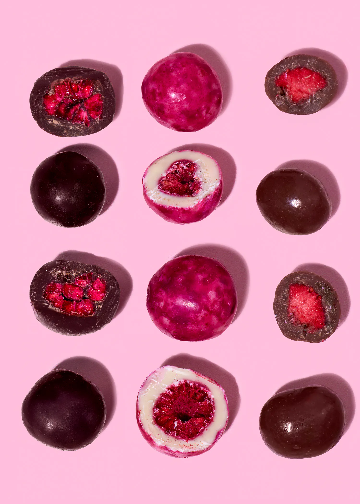 une collection de bonbons enrobés de chocolat noir et de bonbons roses, certains entiers et d'autres coupés en deux révélant un centre de fruits rouges, sur un fond rose uni.