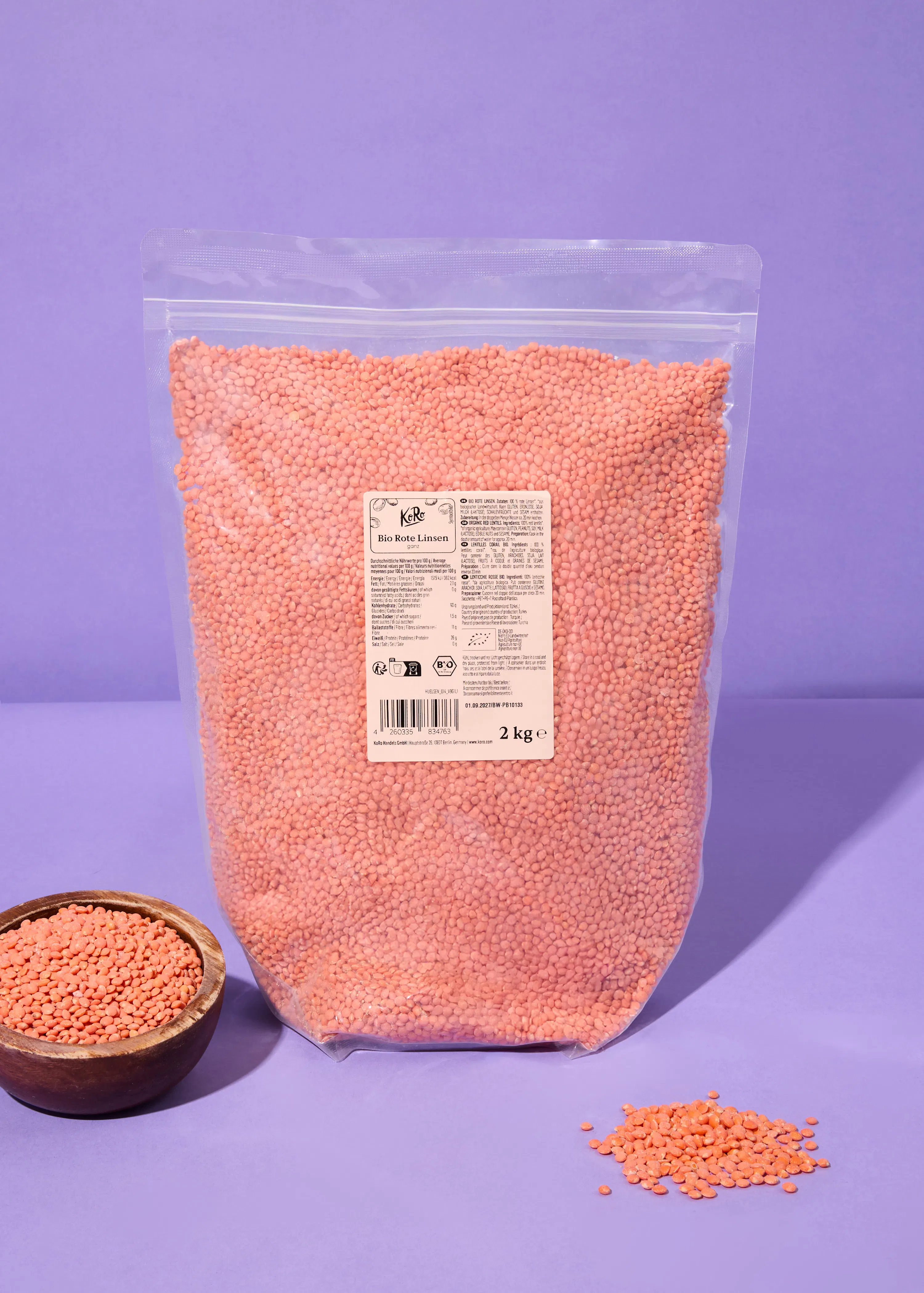 una grande busta trasparente di lenticchie rosse biologiche da 2 kg, una ciotola di legno con lenticchie e alcune lenticchie sparse su una superficie viola.