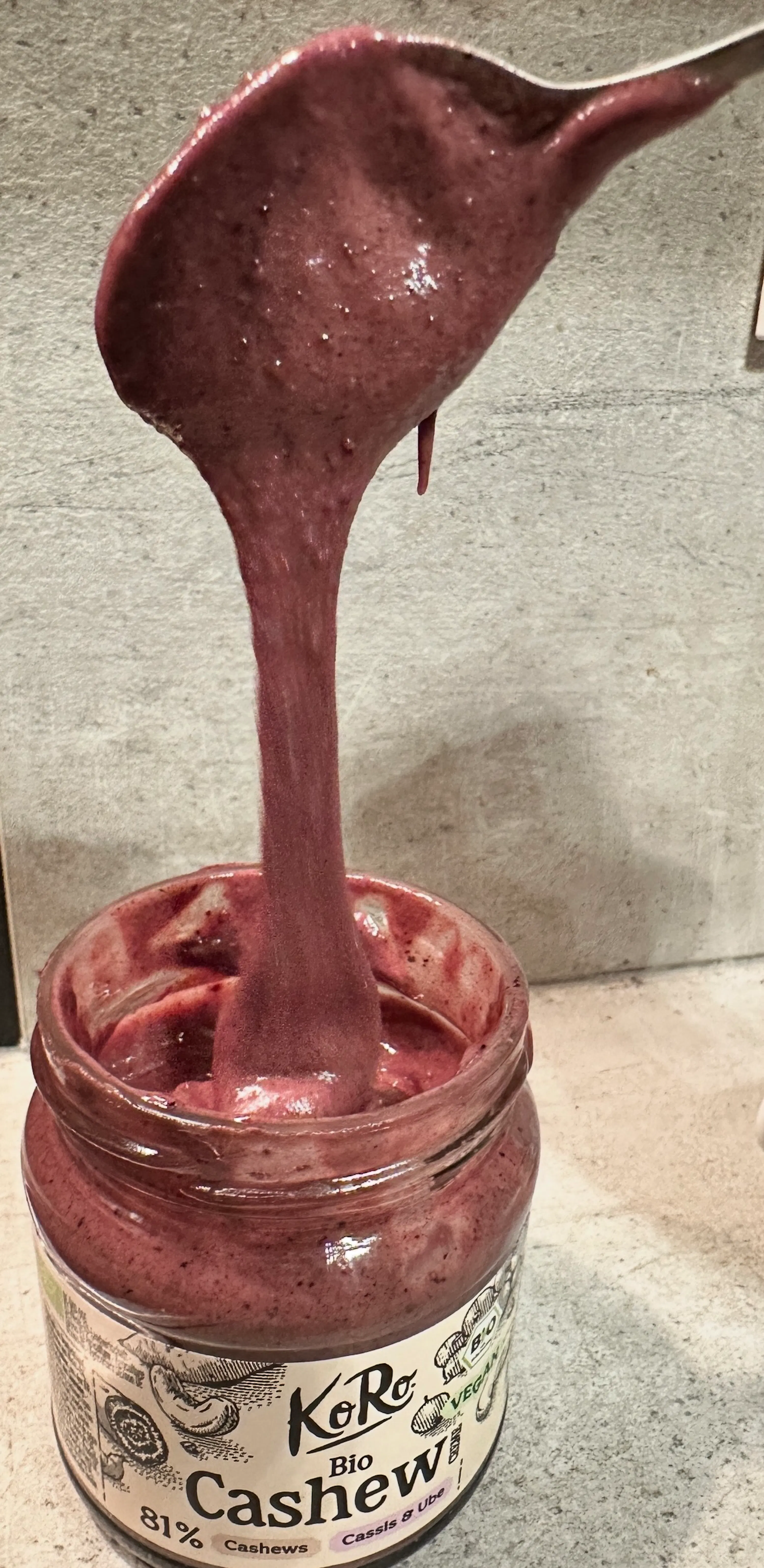 a spoon lifts thick, purple-red koro bio cashew butter with cassis and ube from a jar, the butter stretching and dripping.
