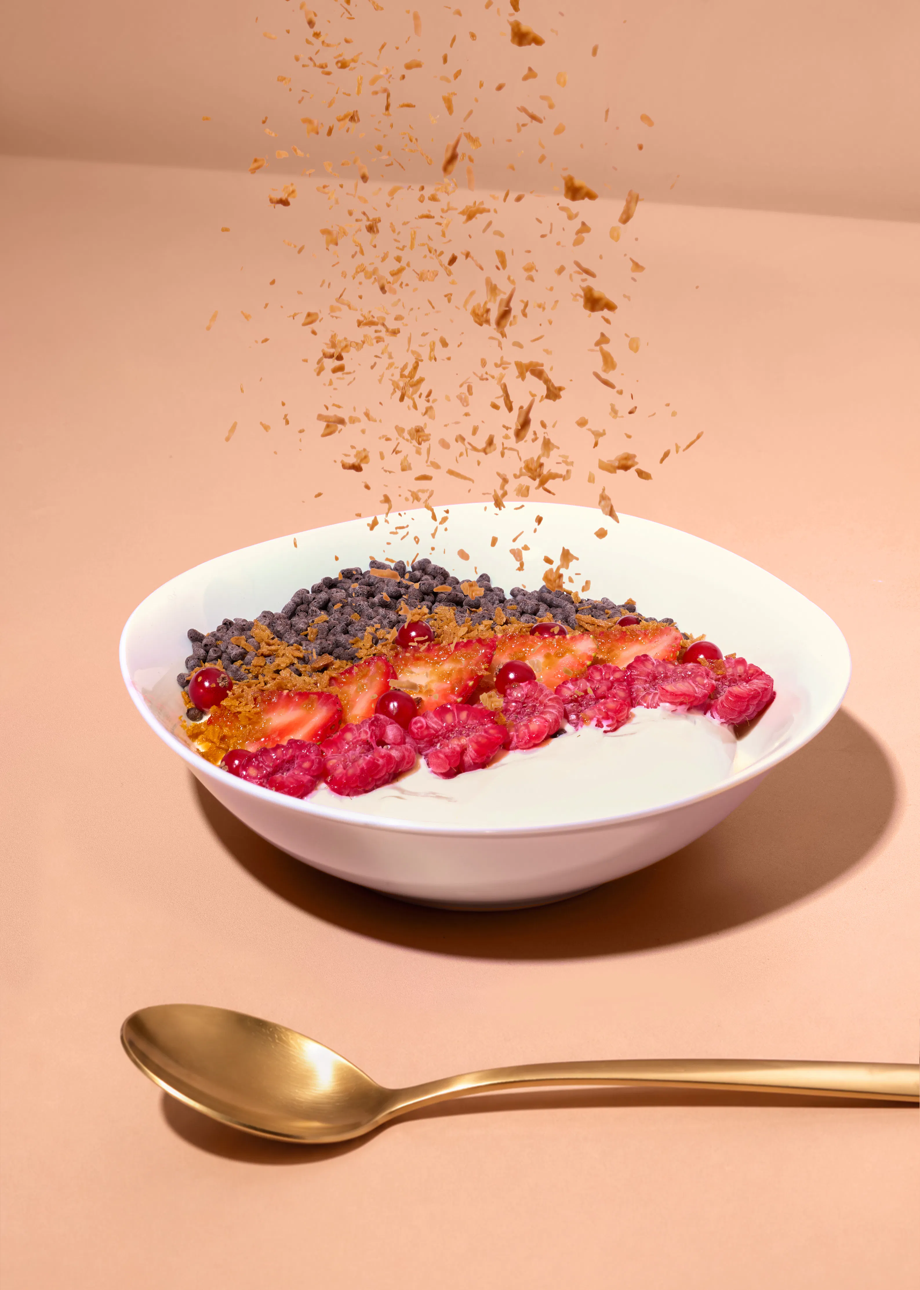 eine weiße schüssel mit joghurt, beeren, schokostückchen und darüber herabfallenden knusprigen flocken vor einem goldenen löffel auf beigem hintergrund.