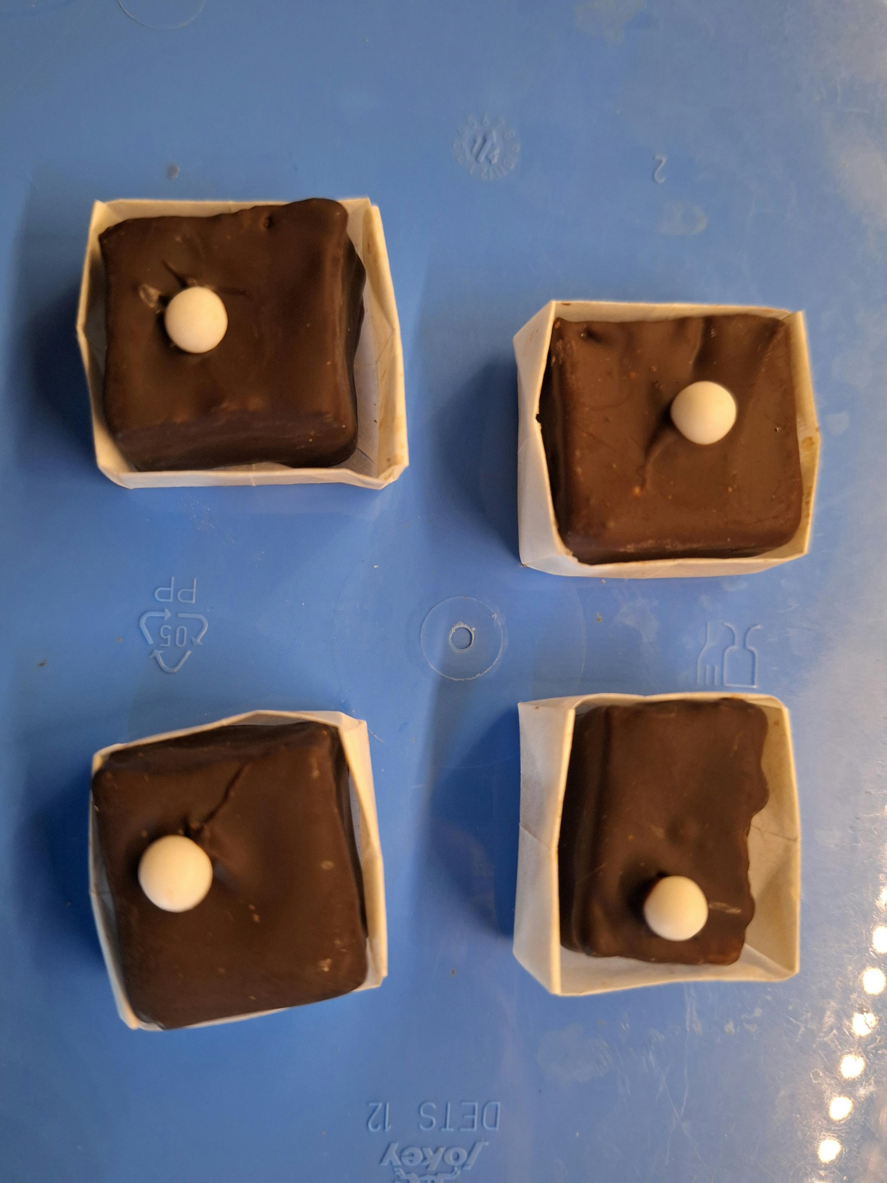 quattro dolcetti quadrati al cioccolato con una pallina bianca sopra, in pirottini di carta su una superficie blu.
