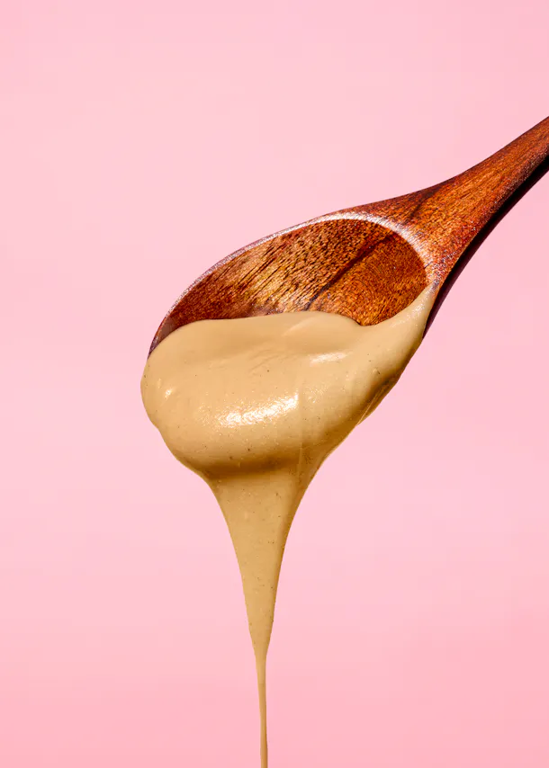 ein holzlöffel mit einer dicken, hellbraunen flüssigkeit, die vor einem rosa hintergrund heruntertropft.