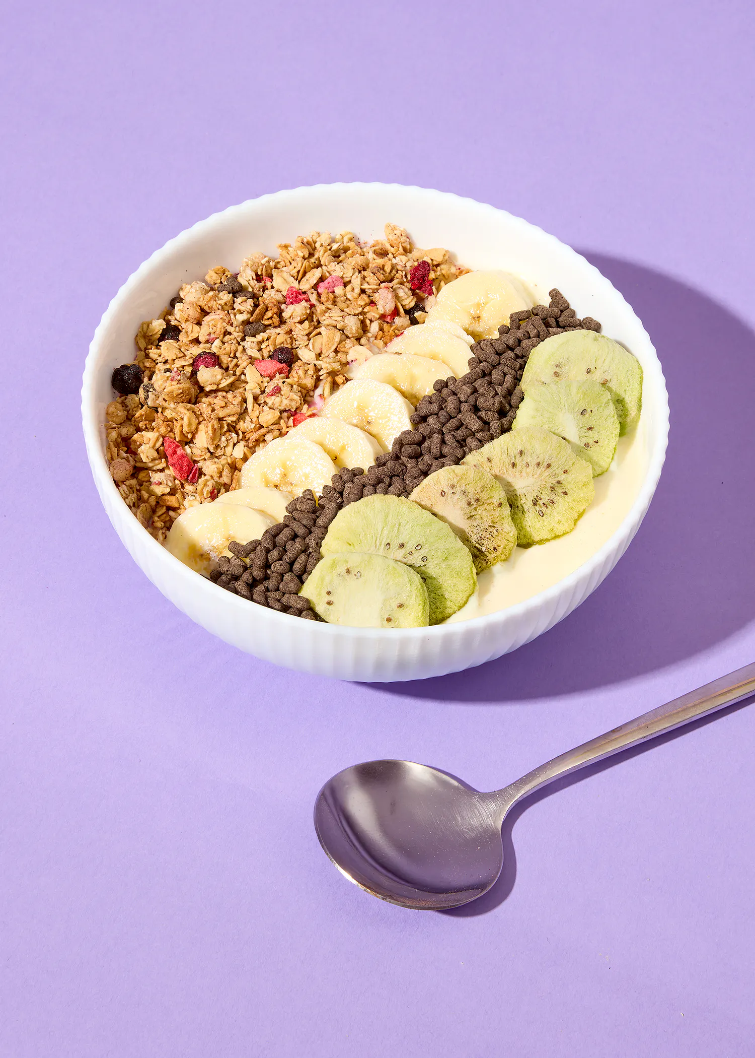 a white bowl filled with diagonal stripes of granola, sliced bananas, chocolate cereal puffs, and dried kiwi slices, resting on a purple surface next to a silver spoon.
