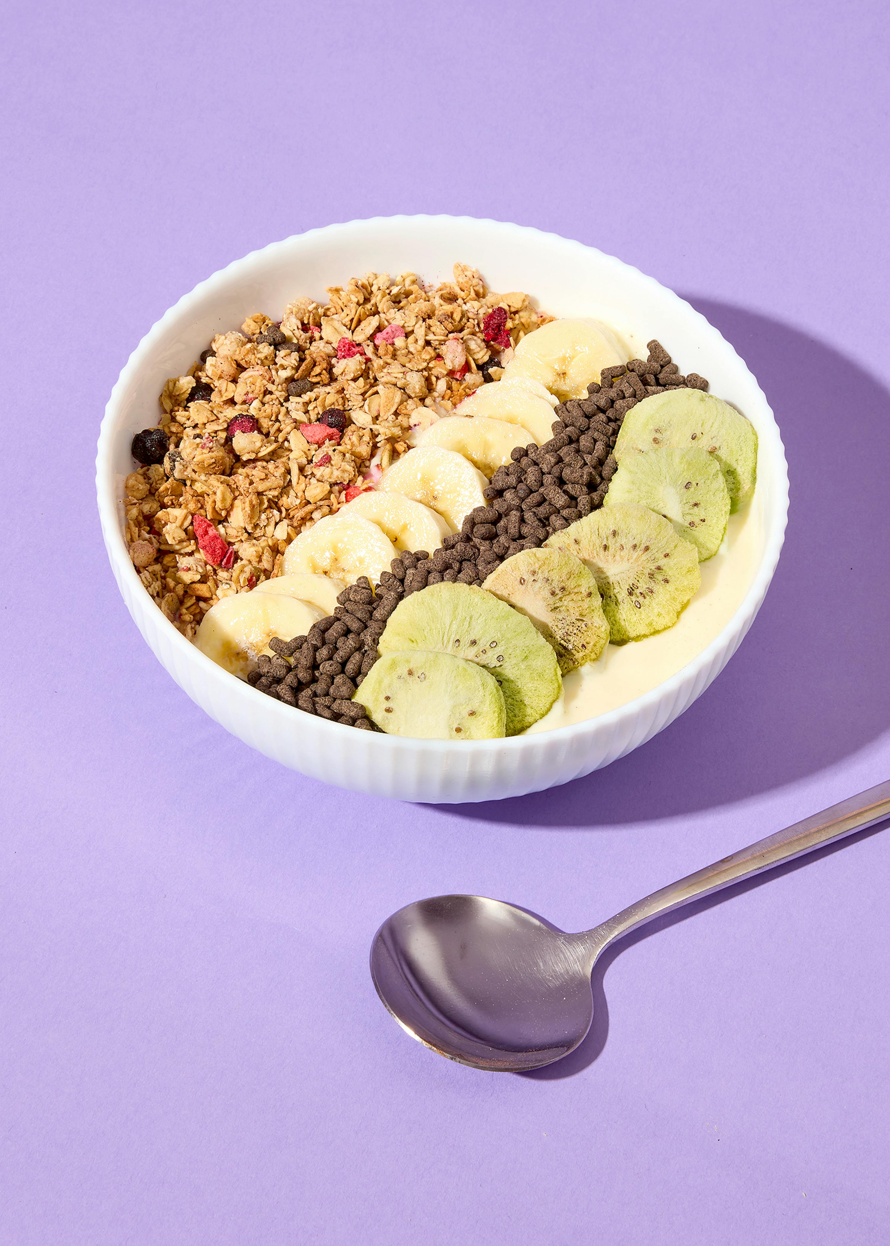 eine weiße schale mit einer frühstücks-bowl, die diagonal mit granola, bananenscheiben, schokoladen-crispies und getrockneten kiwischeiben belegt ist, steht auf einem lila hintergrund neben einem löffel.
