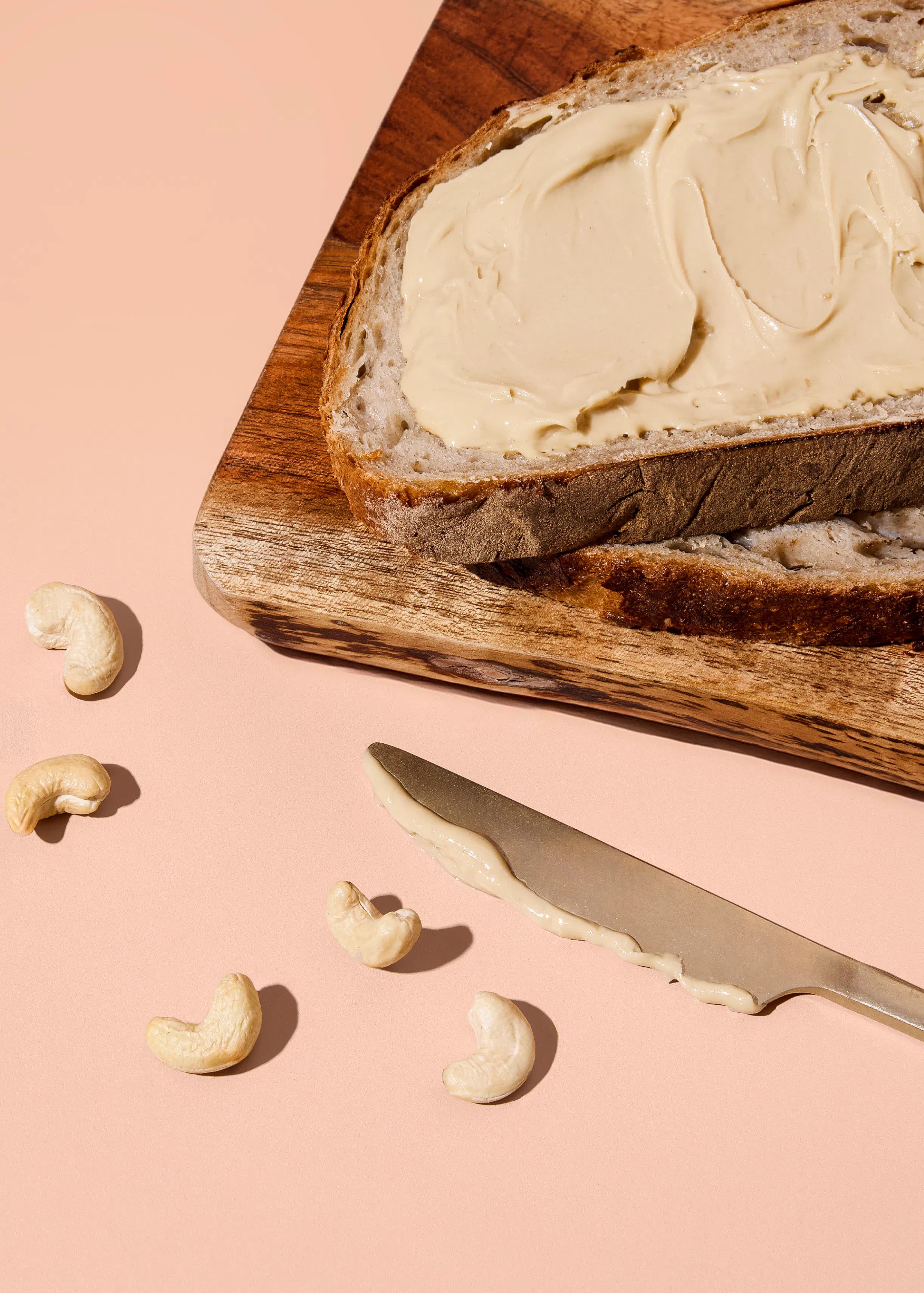 eine scheibe brot mit cashewbutter, ein messer mit cashewbutter und verstreute cashewnüsse liegen auf einem holzbrett vor einem rosa hintergrund.