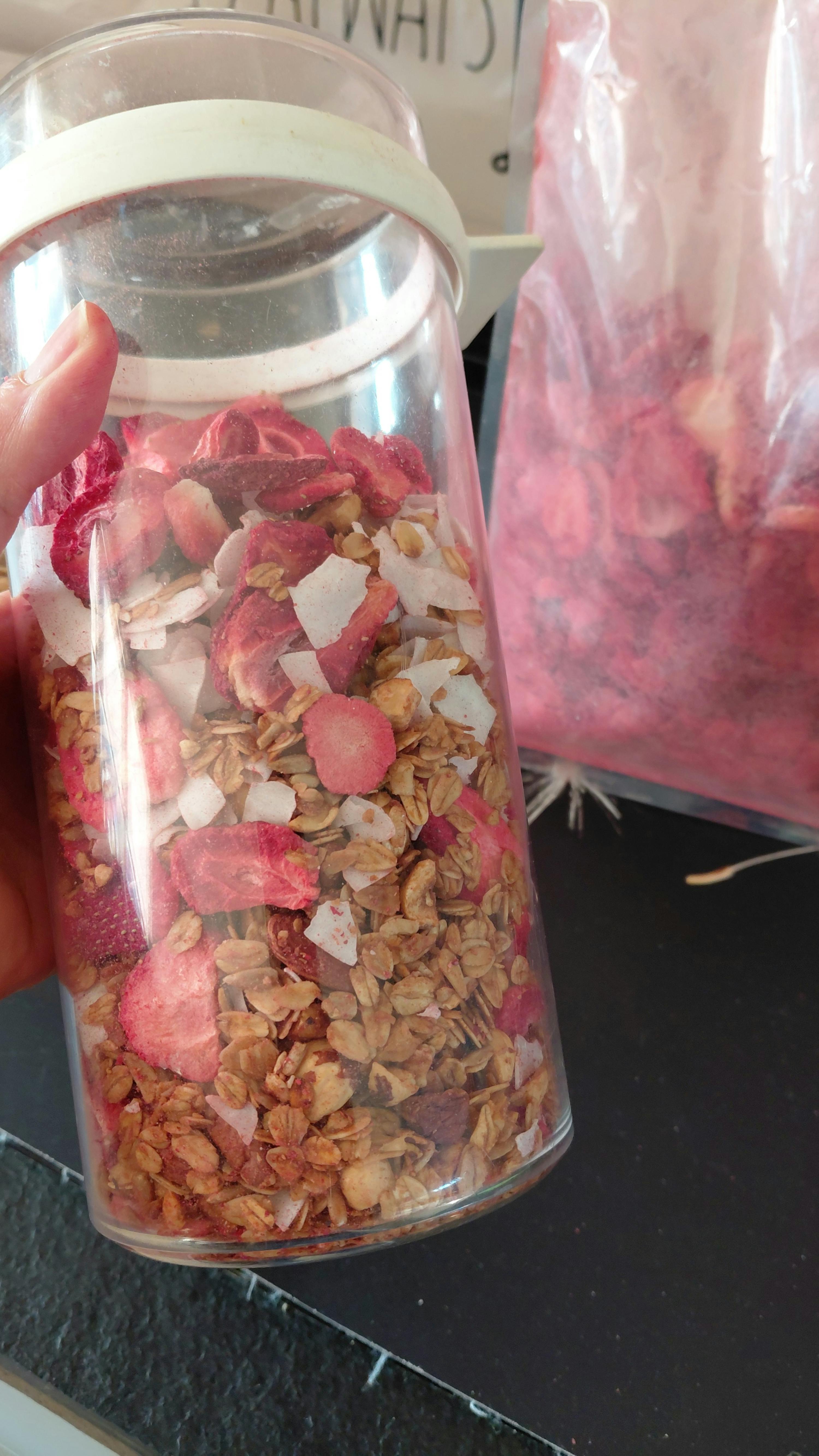 un barattolo trasparente pieno di granola, fragole essiccate e scaglie di cocco, con una busta rosa sfocata sullo sfondo.