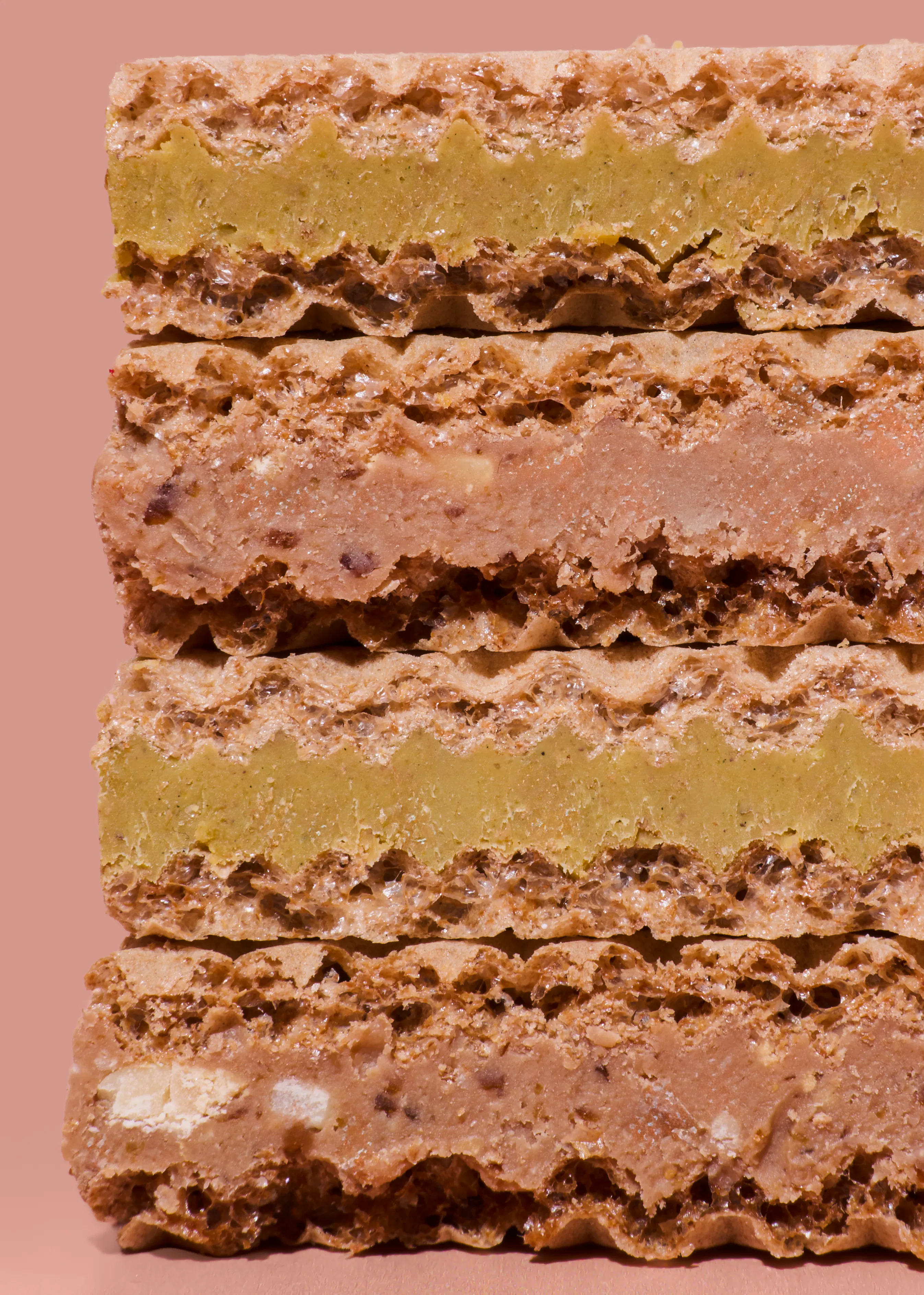 primer plano de galletas de oblea apiladas con capas alternas de relleno verde pistacho y marrón claro con trozos, sobre un fondo rosa pálido.