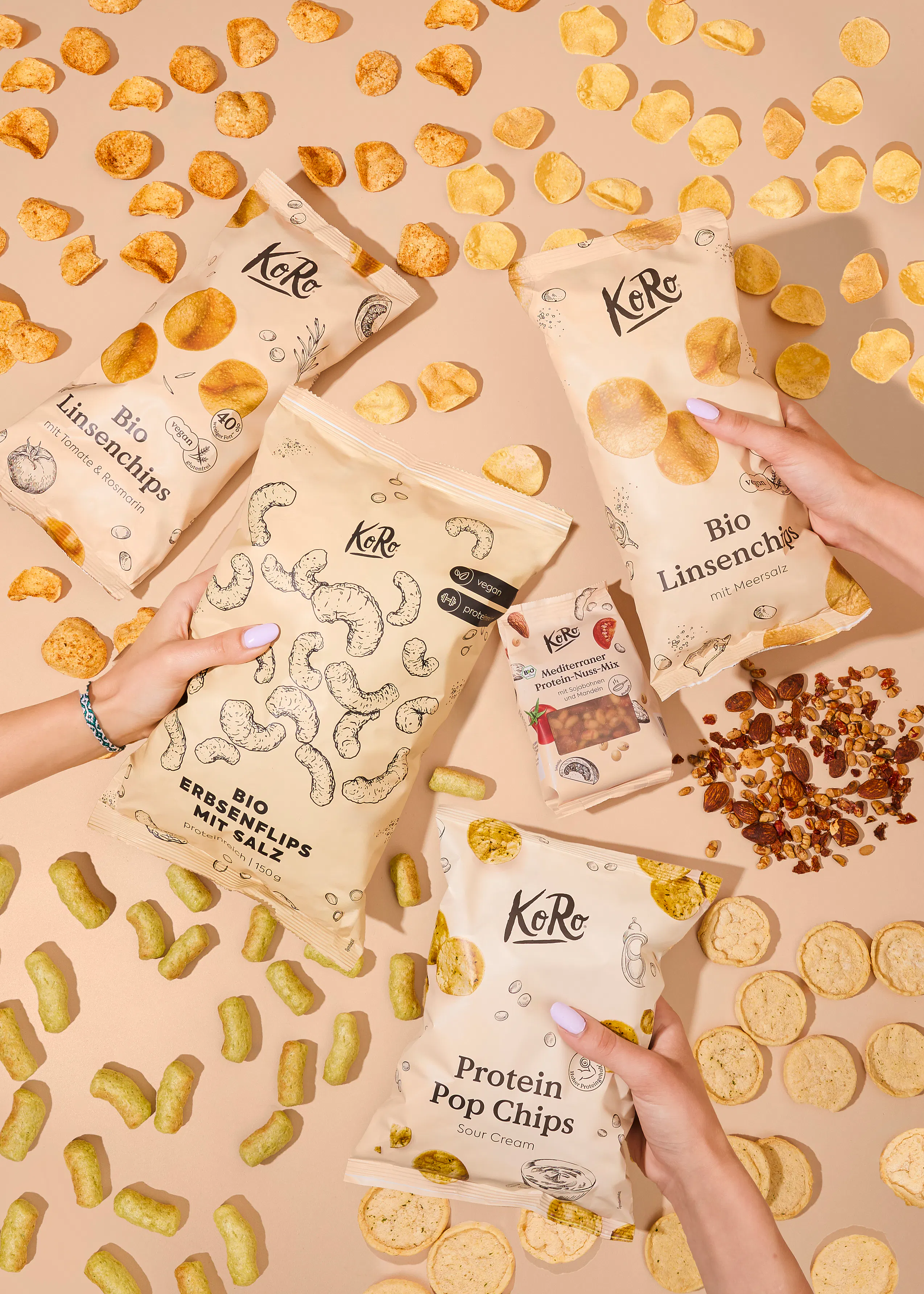 verschiedene packungen koro-snacks, darunter linsenchips, erbsenflips und protein pop chips, liegen auf einem hellen hintergrund, teilweise von händen gehalten und von verstreuten snacks umgeben.