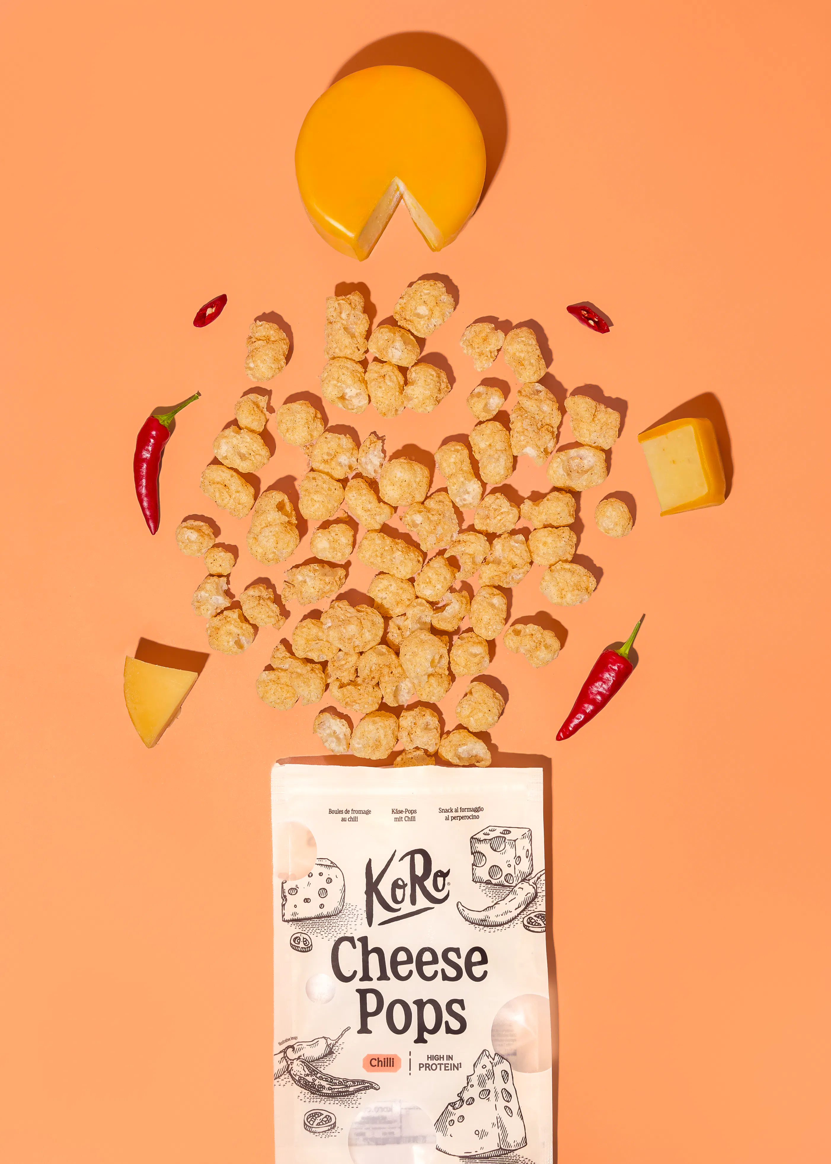 a bag of koro cheese pops with chilli is surrounded by scattered cheese puffs, pieces of cheese, and fresh red chilli peppers on a solid orange background.