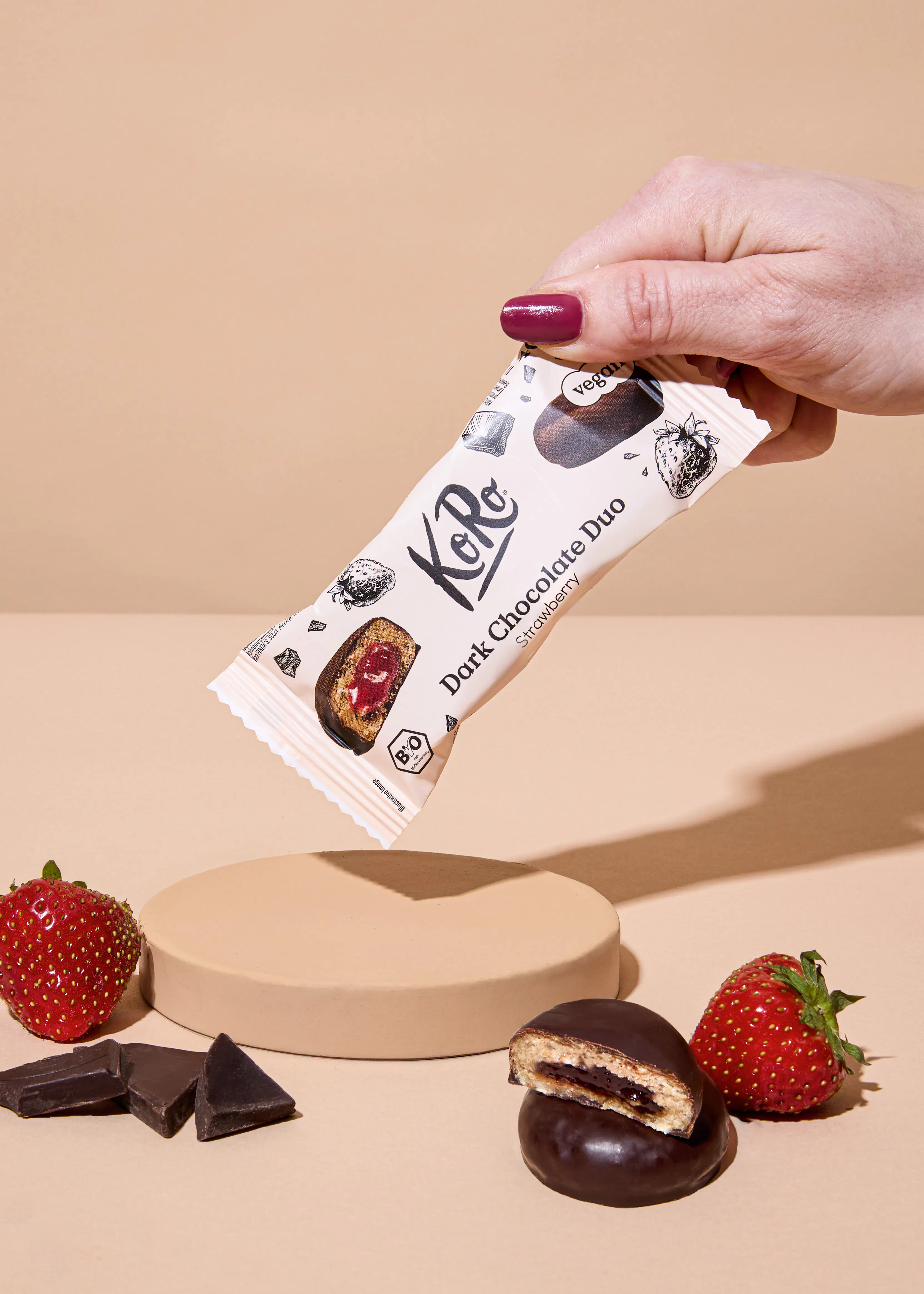 eine hand hält eine koro dark chocolate duo strawberry riegelverpackung, während auf einem beigen tisch frische erdbeeren, schokoladenstücke und ein aufgeschnittener schokoladenkeks mit erdbeerfüllung liegen.