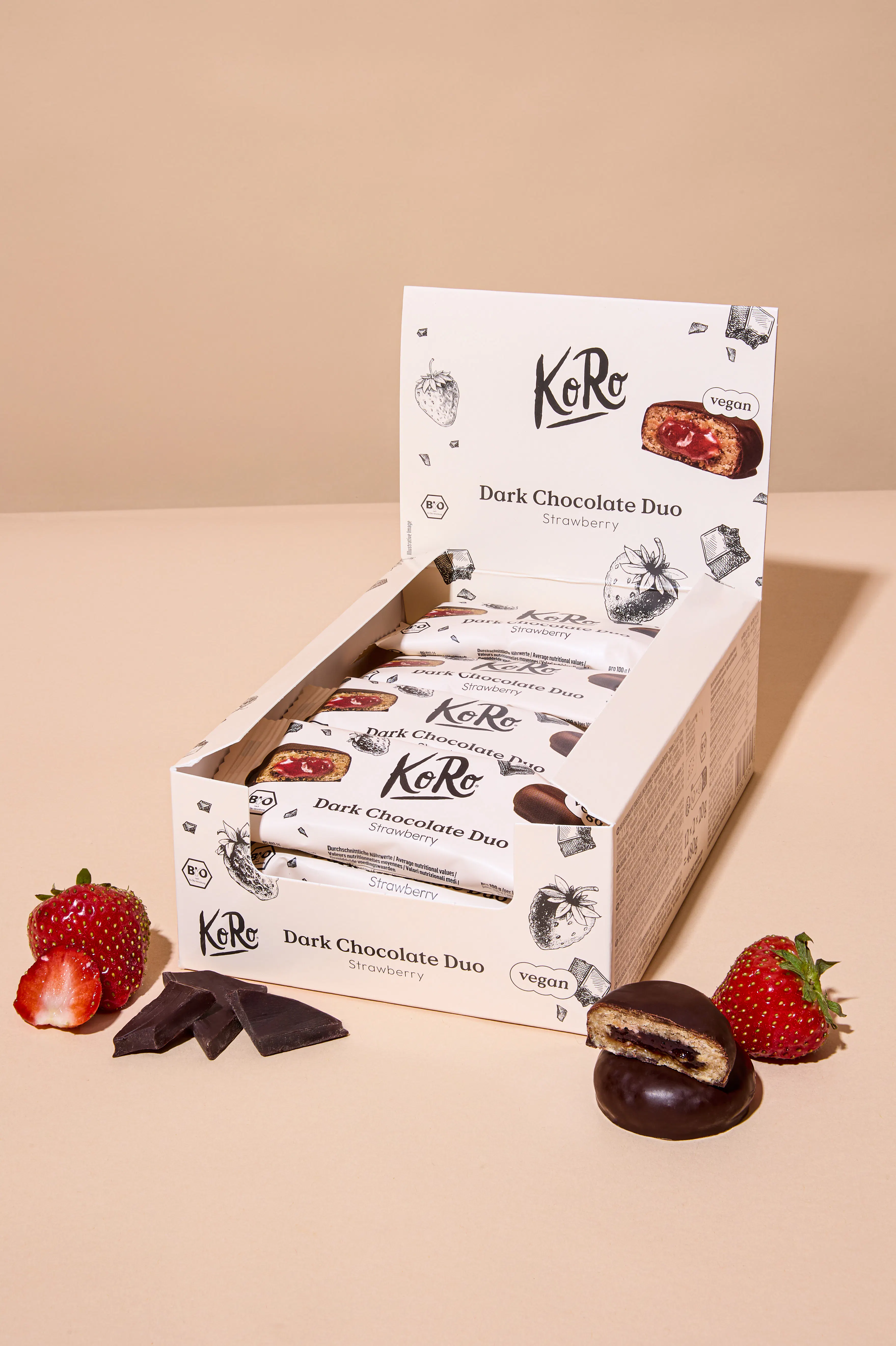 eine offene schachtel mit mehreren einzeln verpackten ko ro dark chocolate duo erdbeer-snacks, umgeben von frischen erdbeeren, schokoladenstücken und einem aufgeschnittenen snack, der die erdbeerfüllung zeigt.