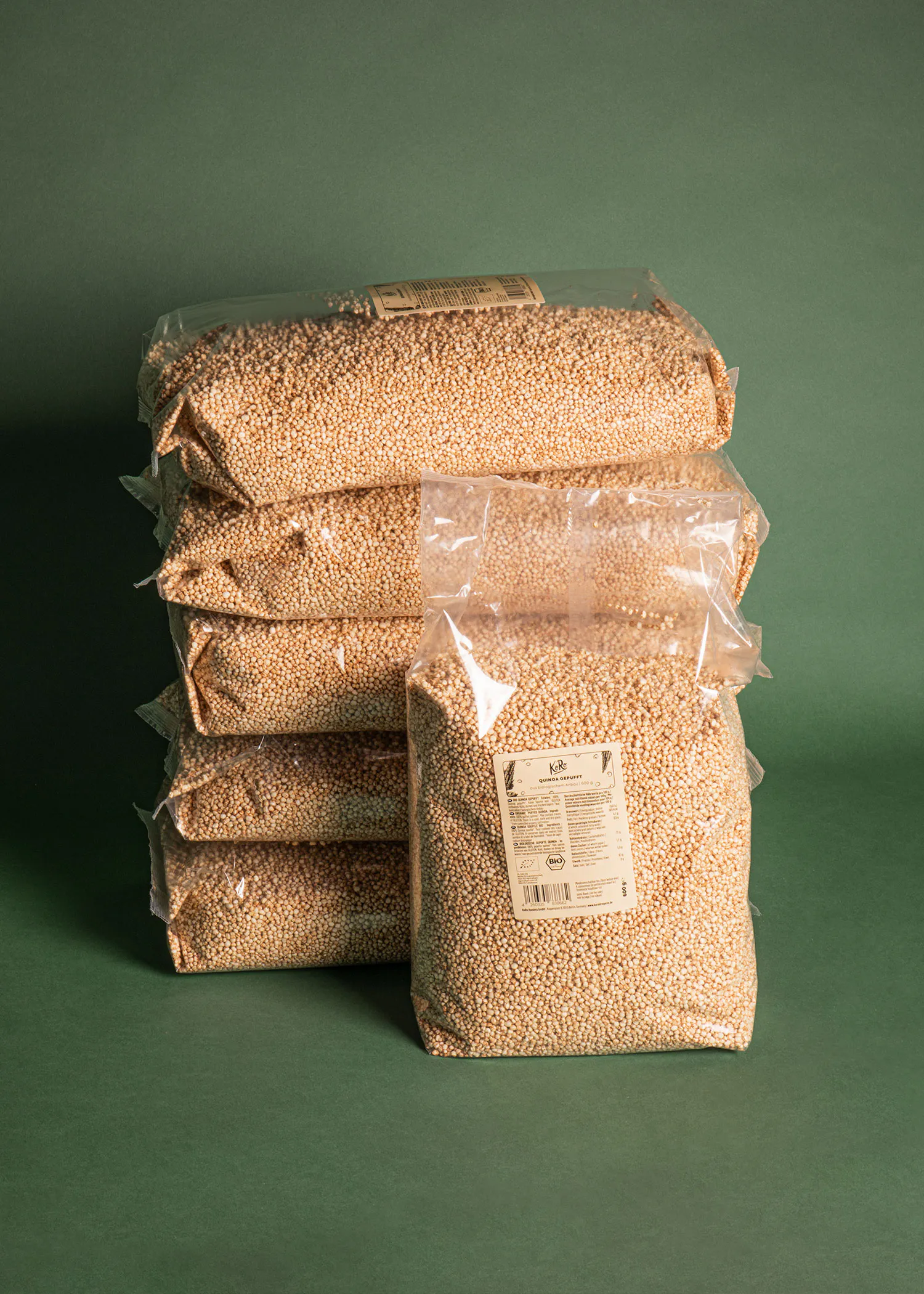 mehrere transparente beutel, gefüllt mit hellbraunem, gepufftem quinoa, sind auf einem dunkelgrünen hintergrund gestapelt.