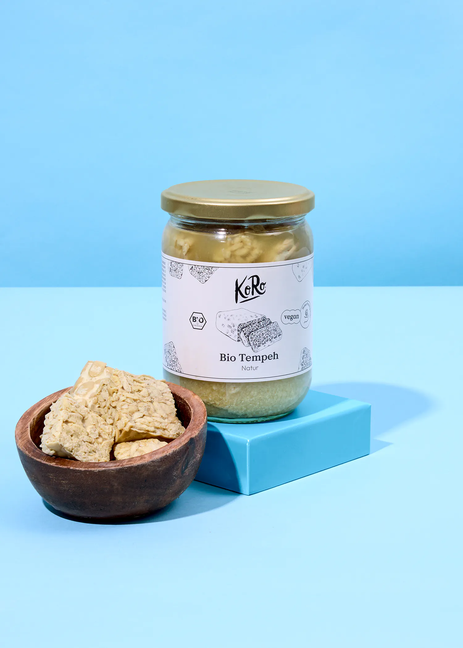 un frasco de tempeh orgánico natural koro sobre un bloque azul y un tazón de madera con trozos de tempeh, ambos sobre un fondo azul claro.