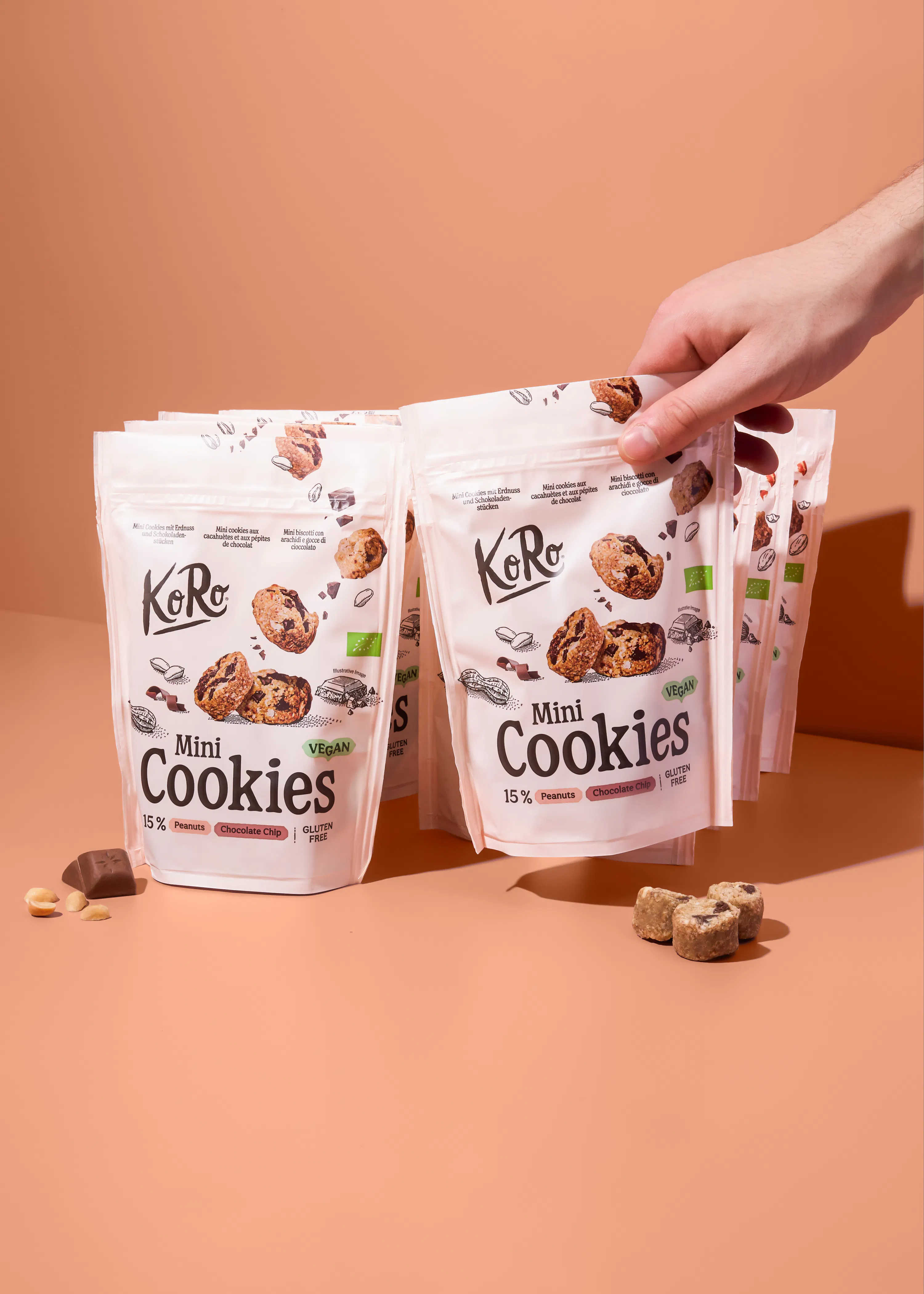 eine hand greift nach einer weißen verpackung von koro mini cookies, die als vegan, glutenfrei, mit 15% erdnüssen und schokoladenstückchen gekennzeichnet sind, während weitere verpackungen und lose zutaten auf einem tisch liegen.