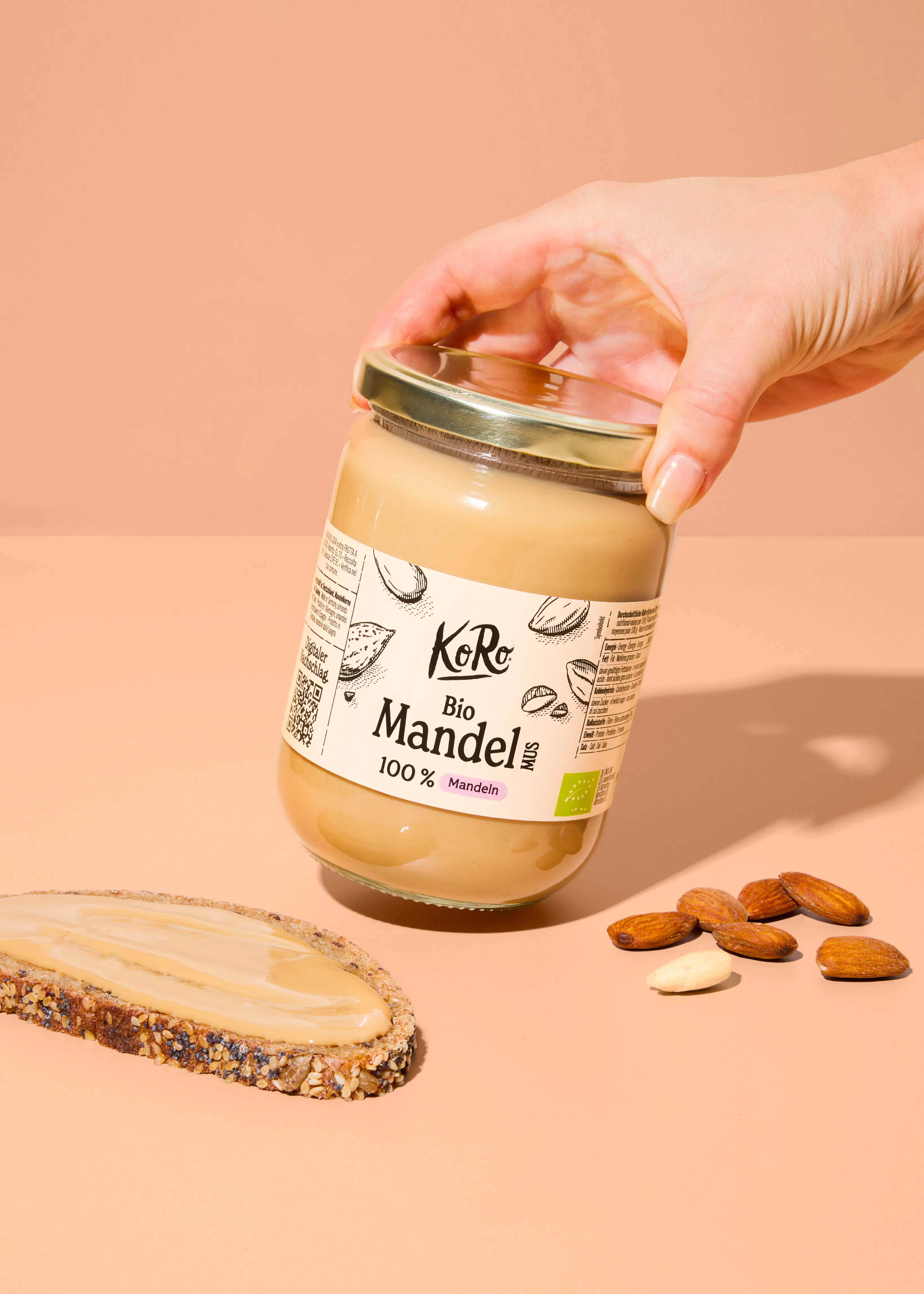 a hand holds a glass jar of koro organic almond butter above a slice of bread spread with the butter and a few scattered almonds on a light peach background.