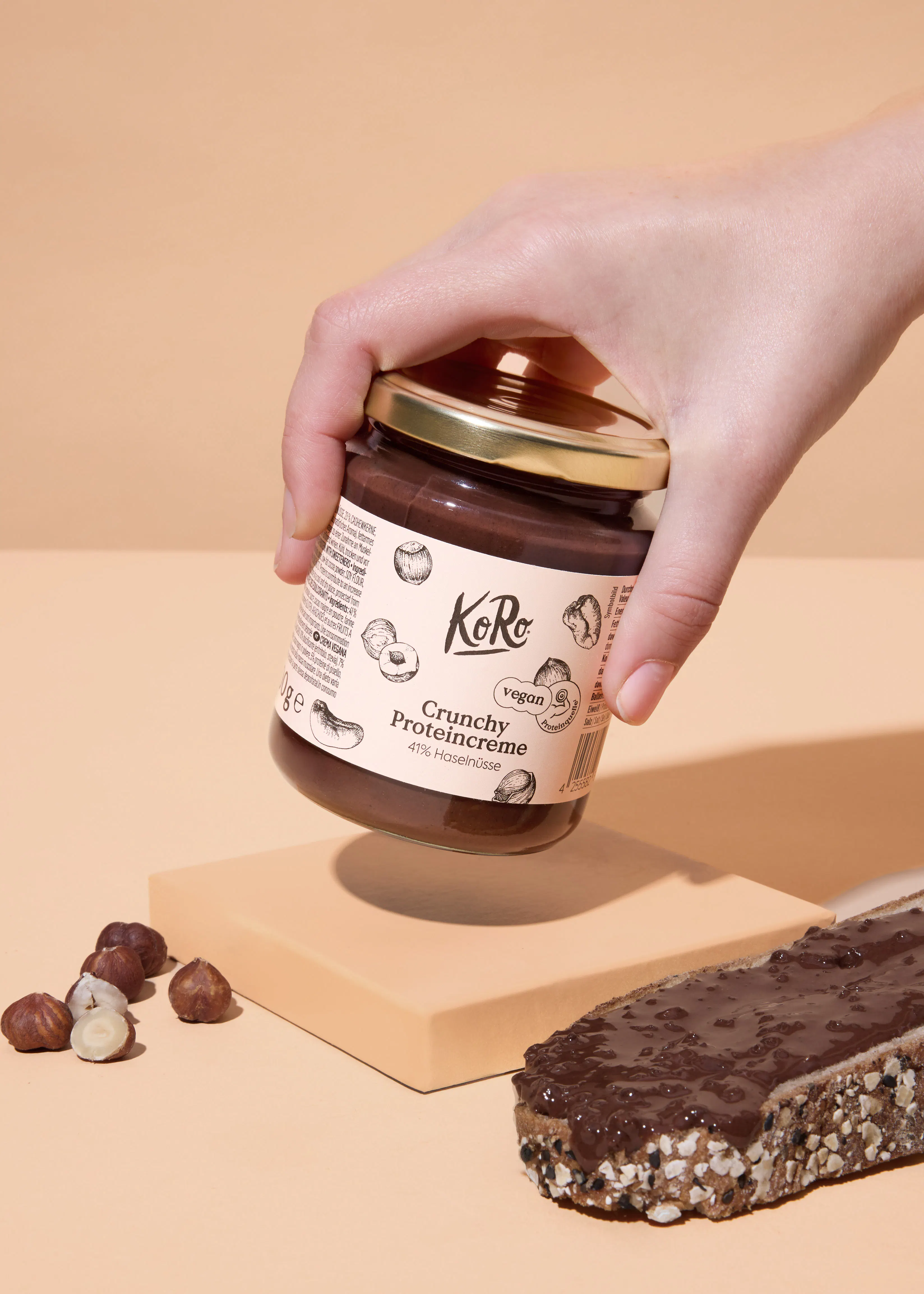 une main tient un pot de crème protéinée croquante aux noisettes koro, avec des noisettes et une tranche de pain tartinée de la même crème en dessous.