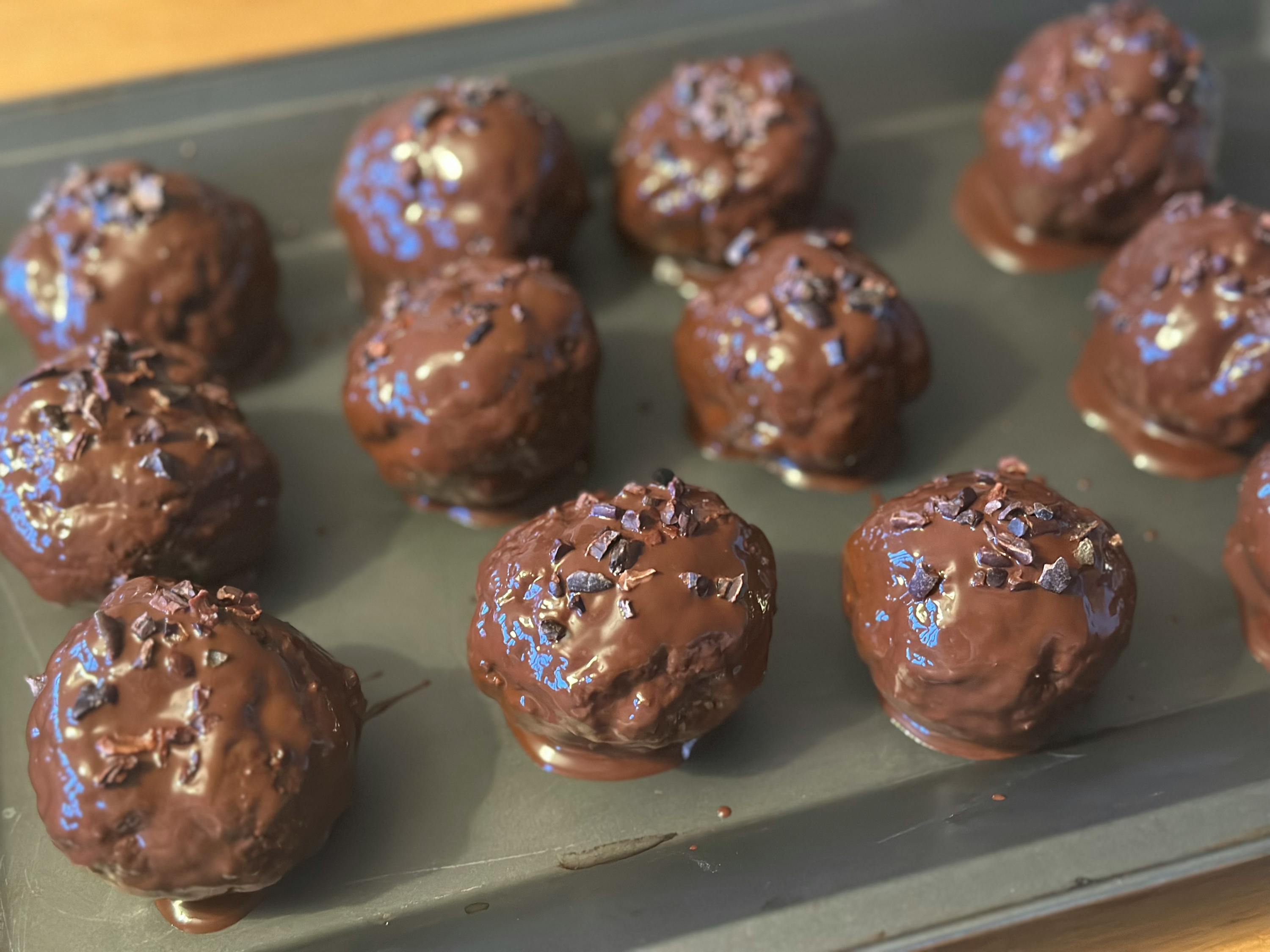 mehrere glänzende, schokoladenüberzogene trüffel, bestreut mit kakaonibs, liegen auf einem dunklen backblech.