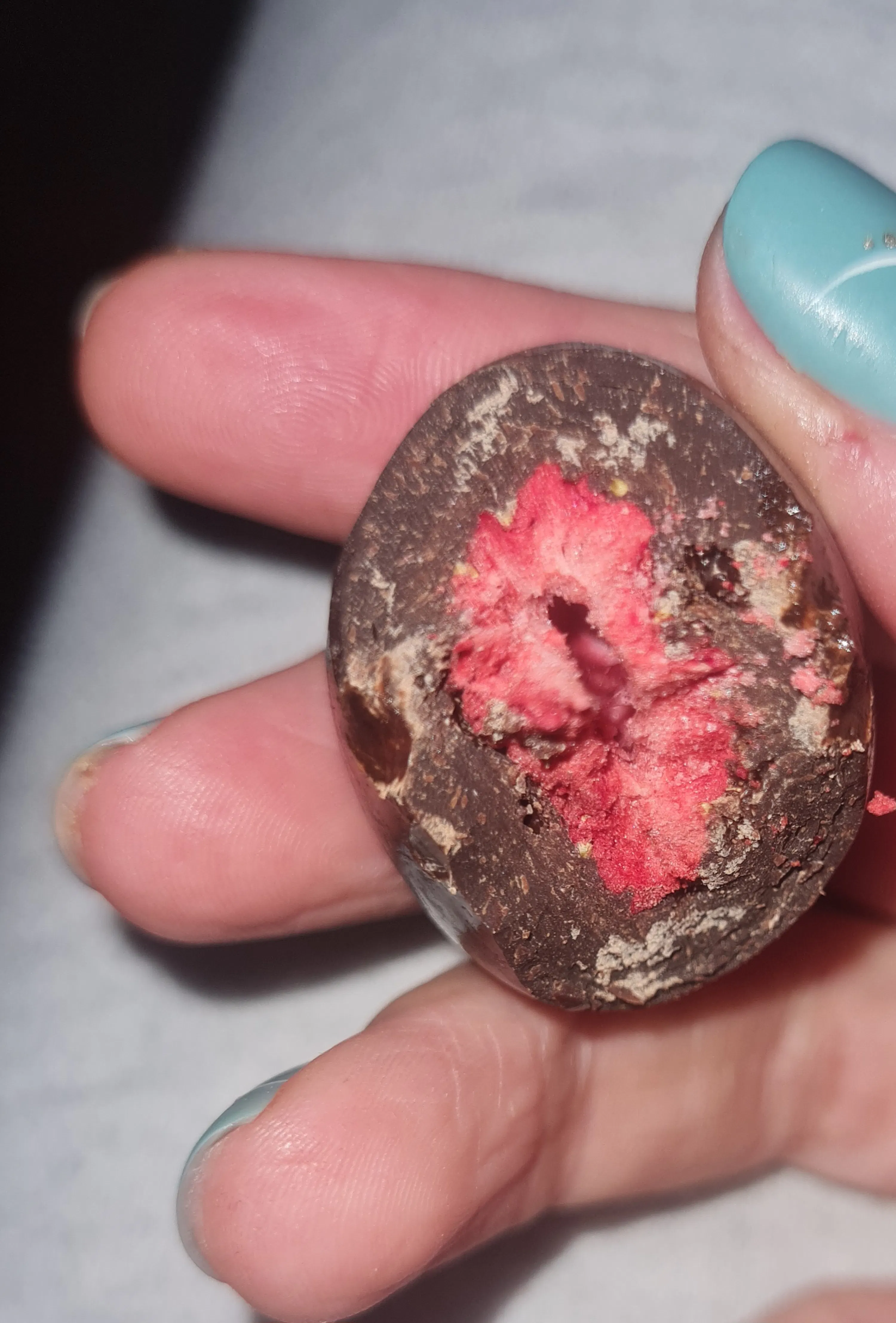 una mano con unghie azzurre tiene un cioccolatino morso che rivela un ripieno rosso brillante, simile a una fragola.
