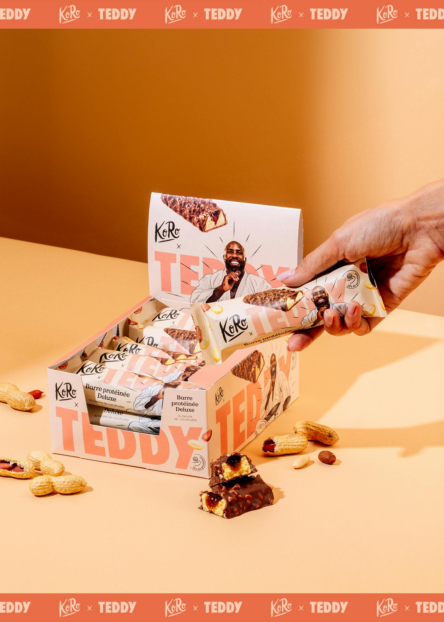 une main prend une barre protéinée deluxe ko ro x teddy au beurre de cacahuète d'une boîte de présentation, avec des cacahuètes et une barre cassée sur une surface orange.