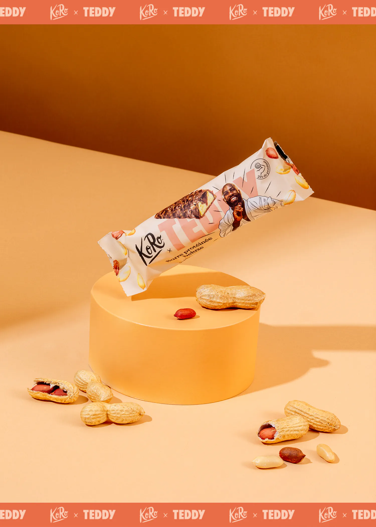 une barre protéinée deluxe ko ro x teddy, avec un homme souriant et une coupe transversale de la barre, flotte au-dessus d'un piédestal orange, entourée de cacahuètes sur un fond orange.