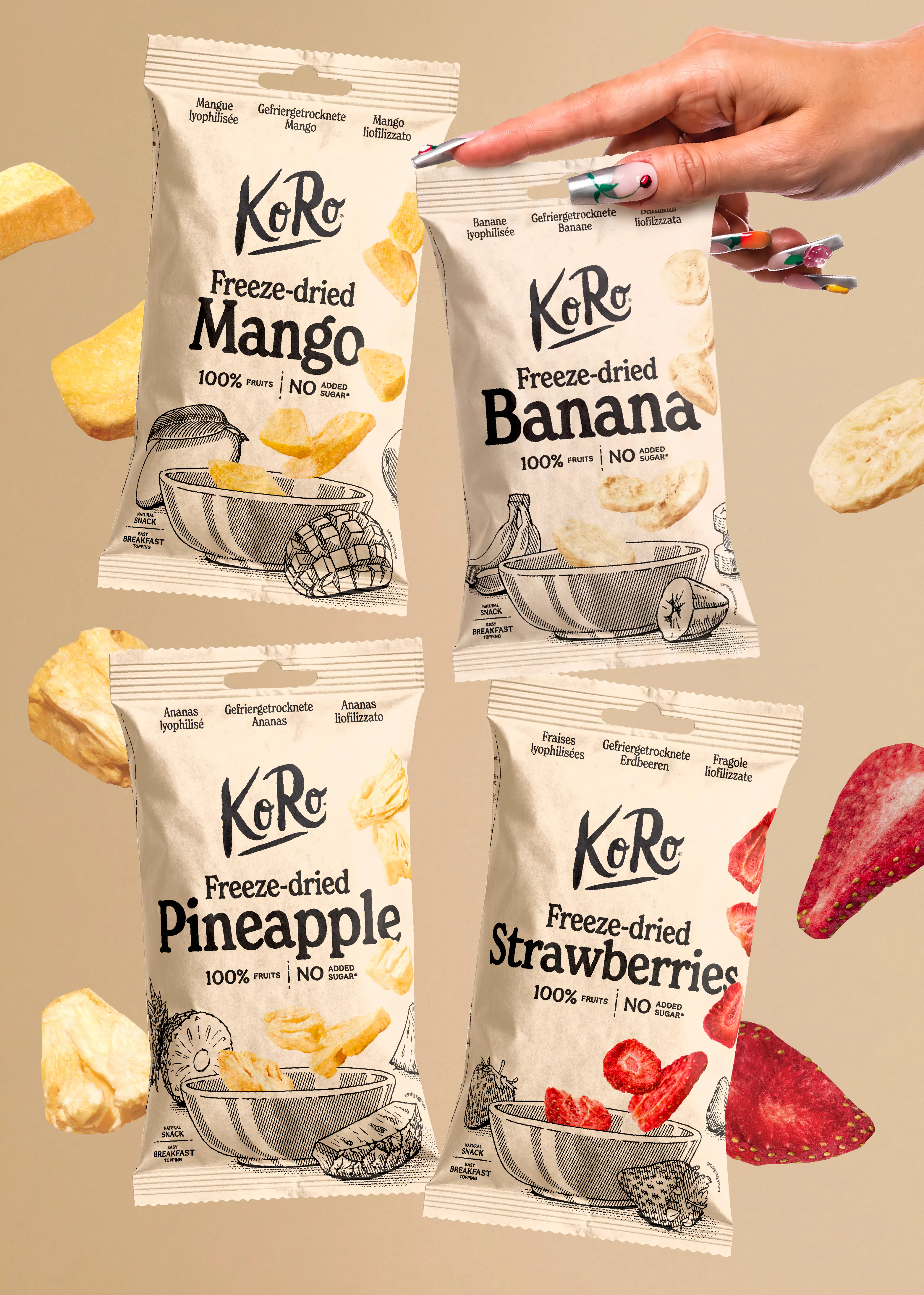 une main tient un sachet de bananes lyophilisées koro au-dessus de trois autres sachets de fruits lyophilisés de la même marque, entourés de morceaux de fruits flottants sur un fond beige.
