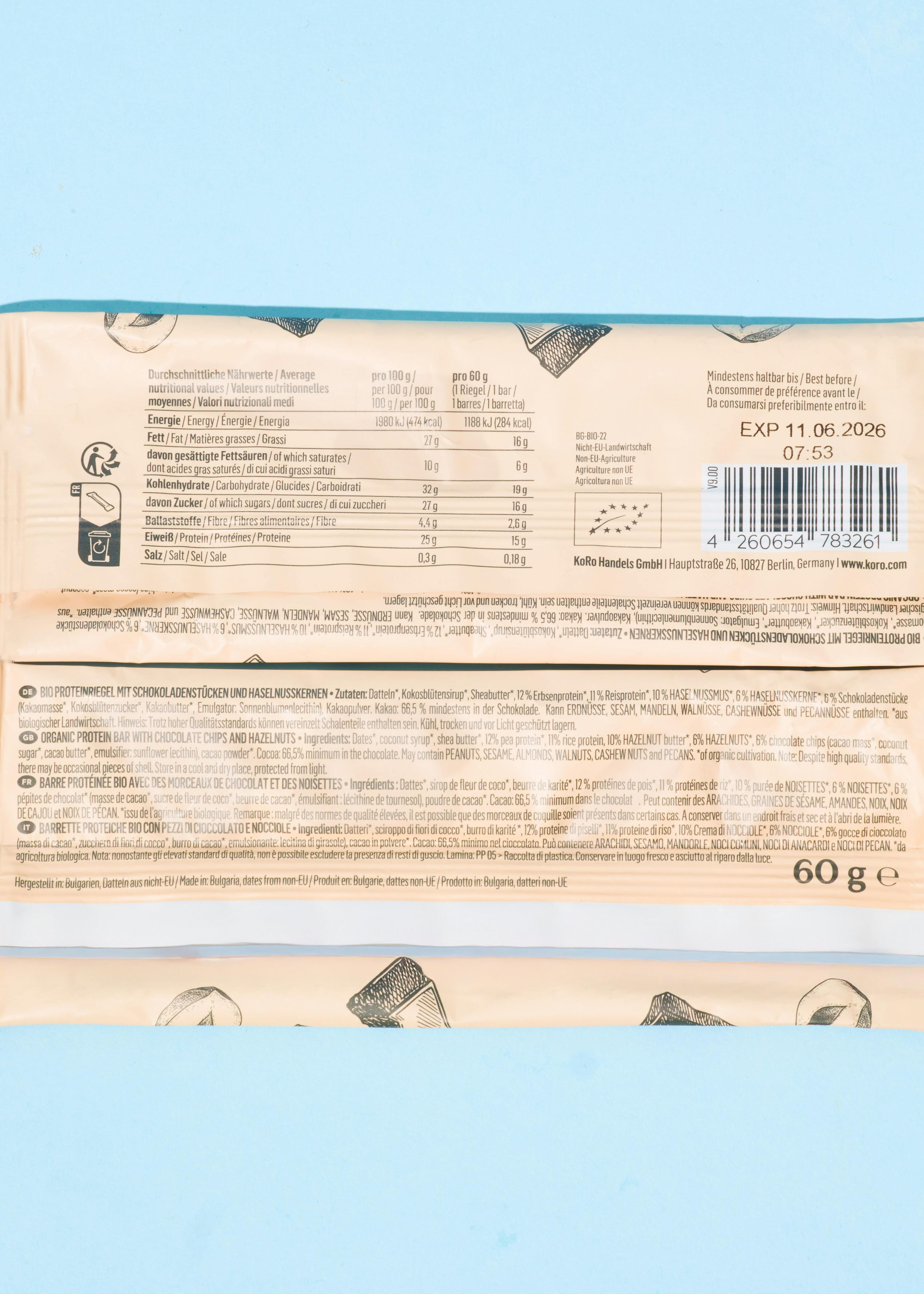 ein bio-proteinriegel mit schokoladenstücken und haselnusskernen, der nährwertangaben, zutatenlisten und einen barcode auf der verpackung zeigt.