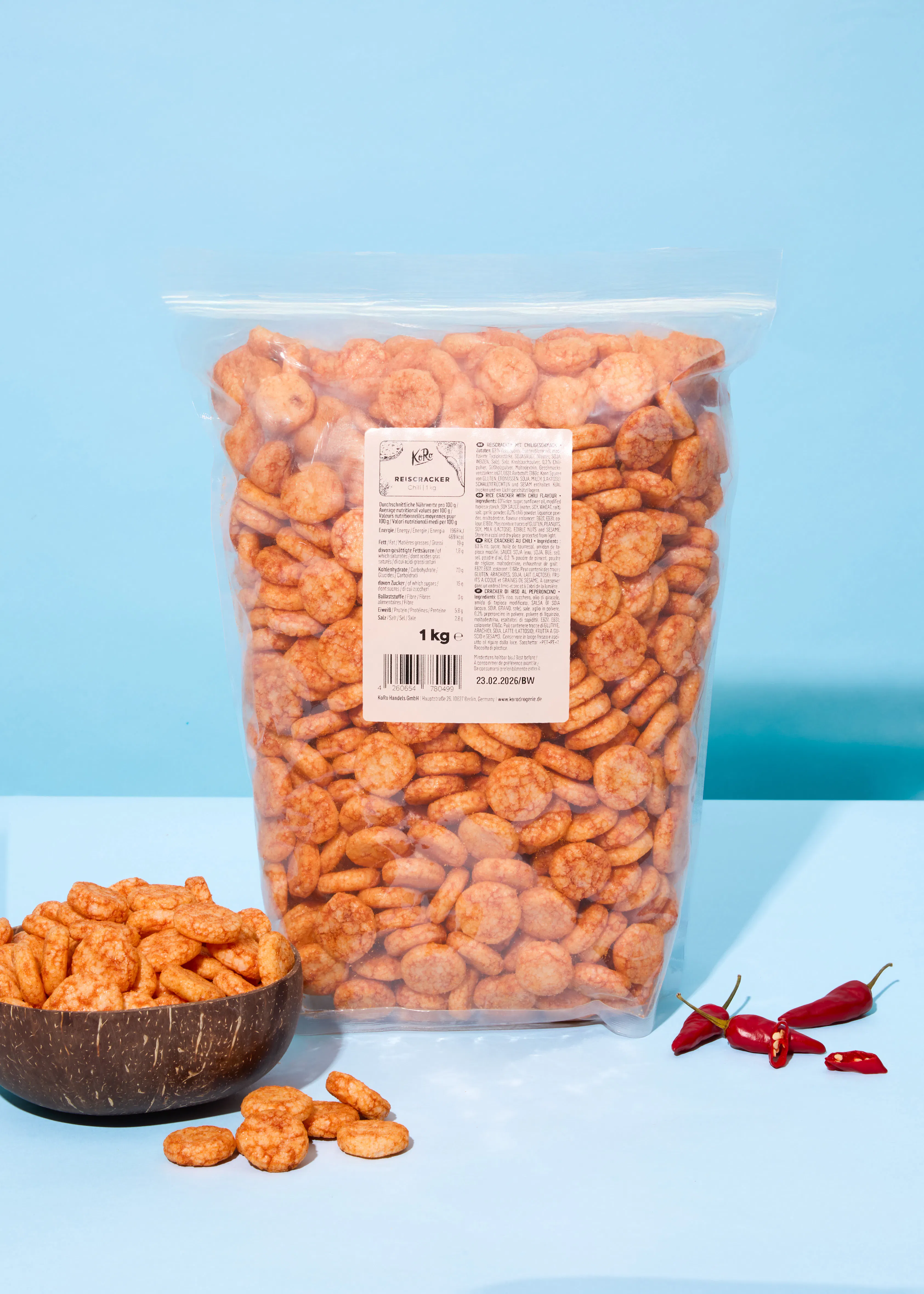 un grand sac transparent de crackers de riz épicés, un bol en bois rempli de crackers et des piments rouges sur une surface bleue.