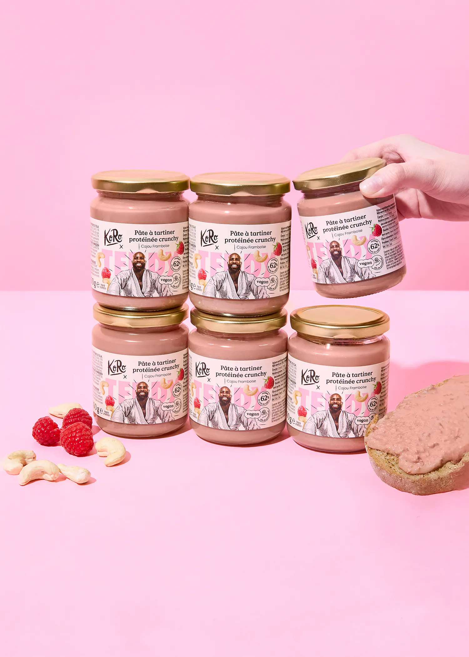 mehrere gläser ko ro crunchy protein-brotaufstrich mit cashew und himbeere, umgeben von frischen himbeeren und cashewnüssen, wobei ein glas von einer hand gehalten wird und ein brot mit dem aufstrich daneben liegt, alles auf einem pinken hintergrund.