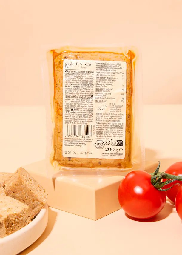 un paquet de tofu bio koro rosso aux tomates séchées et épices est présenté sur un support beige, accompagné de tomates cerises fraîches et d'un morceau de tofu coupé.