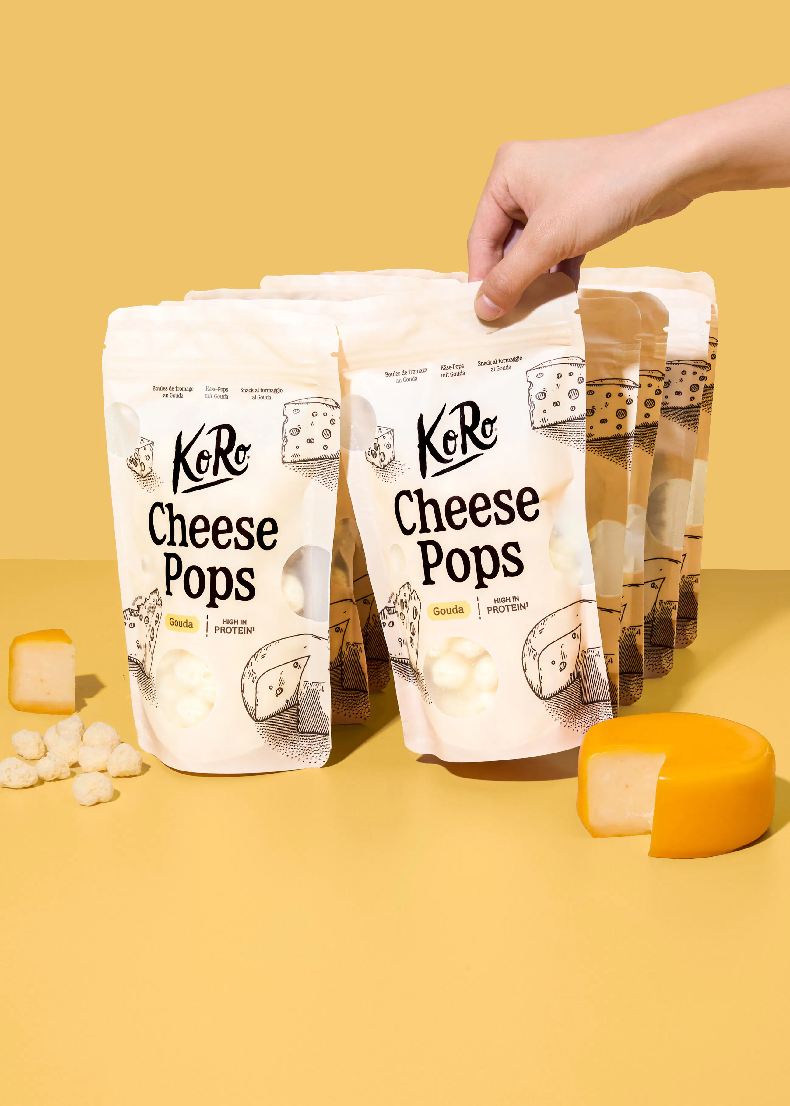 eine hand hält eine packung koro cheese pops vor weiteren packungen auf einem gelben hintergrund, wobei daneben ein stück gouda und einzelne käsebällchen liegen.